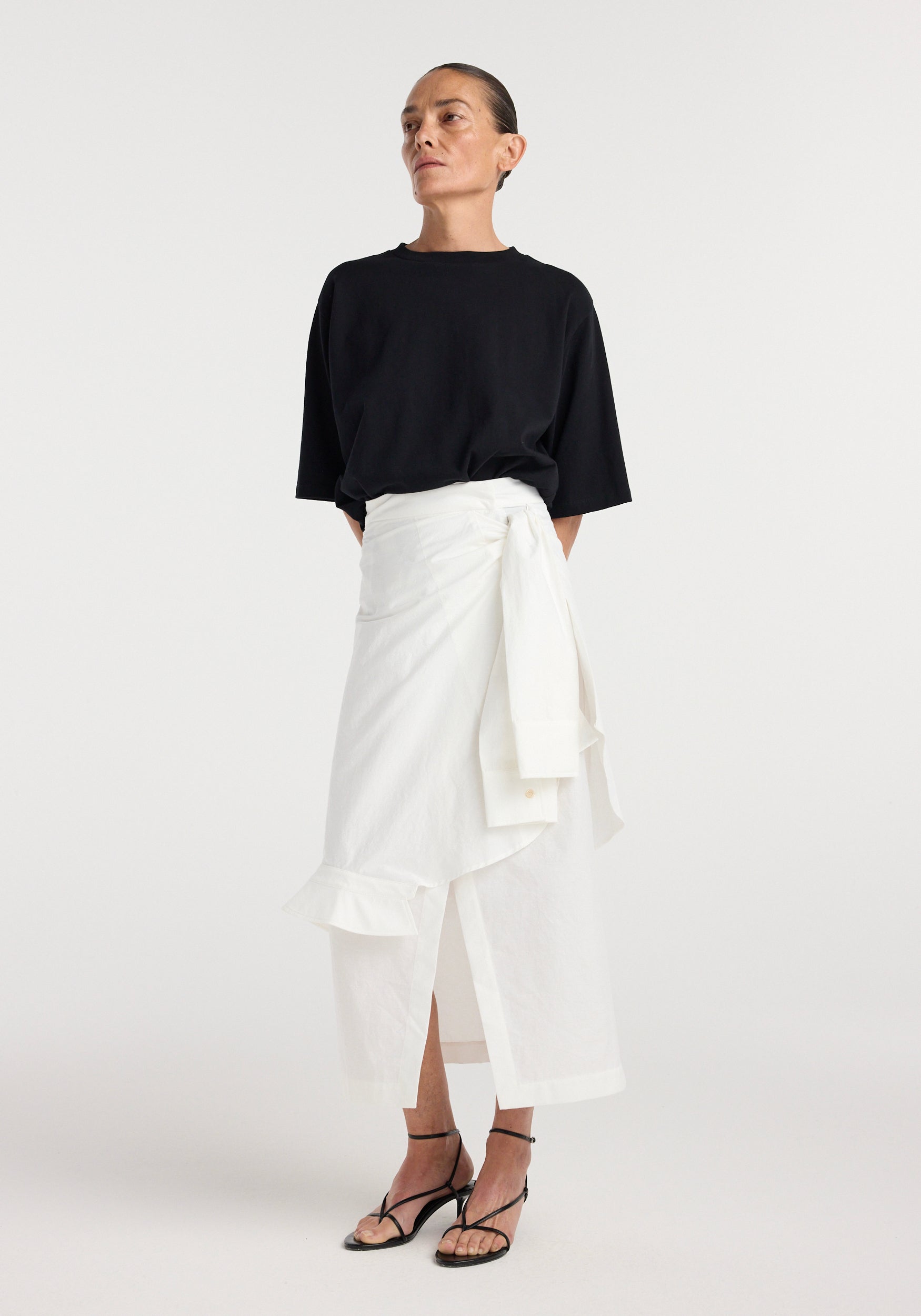 Woman wearing a black oversized t-shirt paired with a white shirt-wrap detailed skirt | optic white, styled with black strappy heeled sandals against a plain background.
