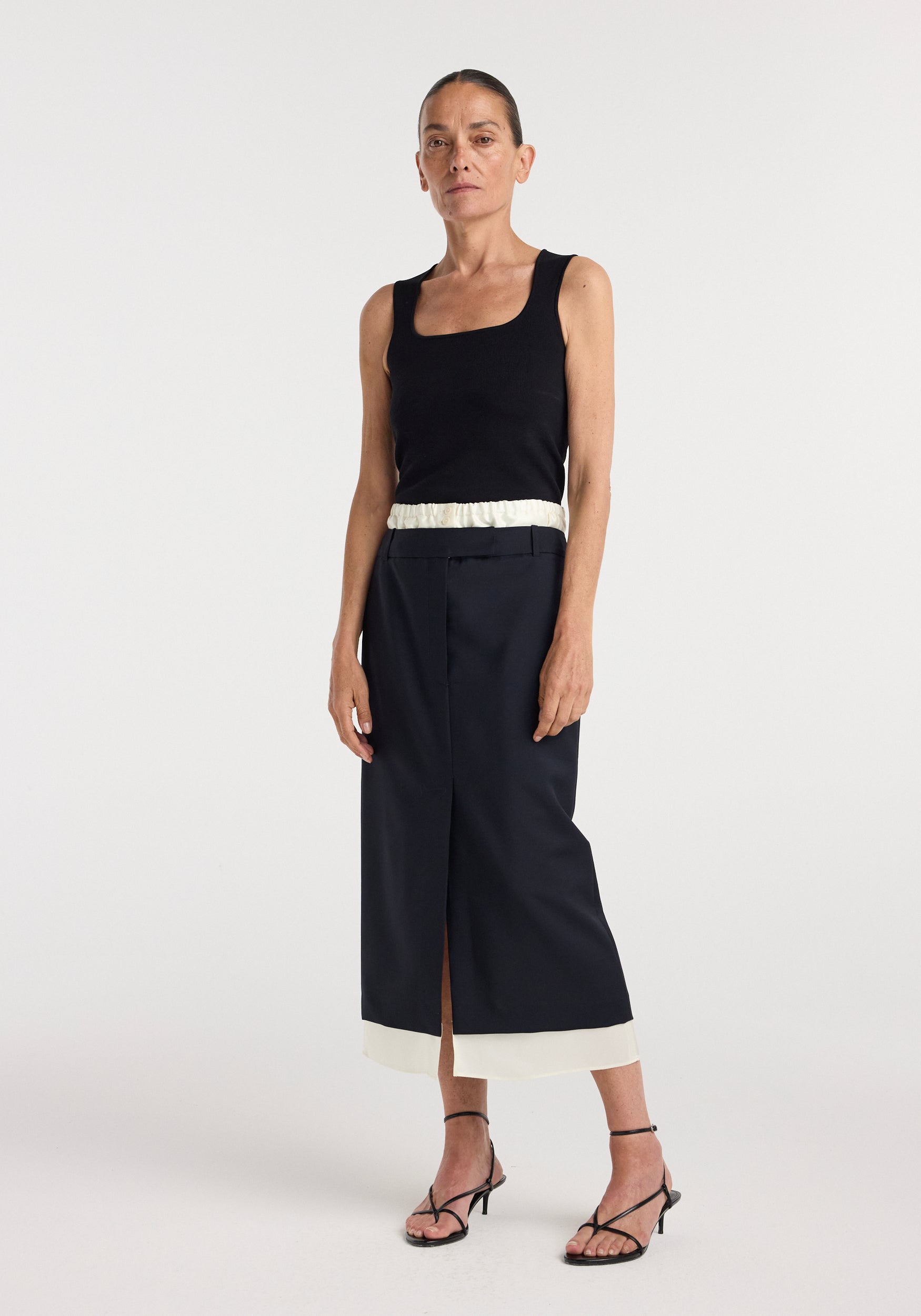 Woman wearing a sleeveless black top paired with a navy Boxer detailed skirt | navy featuring a cream waistband and hem, standing against a plain white background.