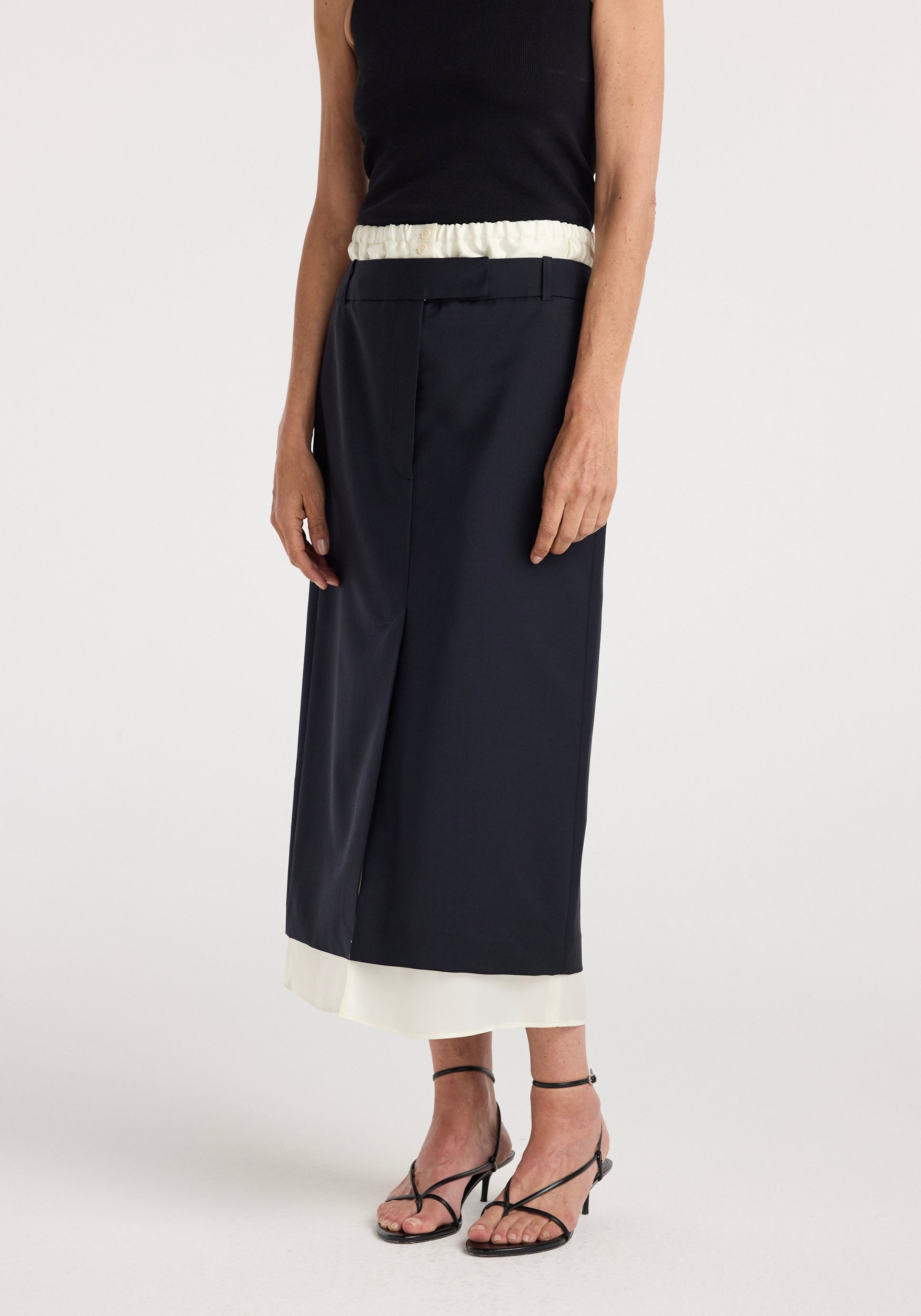 Model wearing a navy Boxer detailed skirt | navy featuring a straight cut with a subtle overlay and a contrasting white hem, paired with black strappy heels and a sleeveless black top.