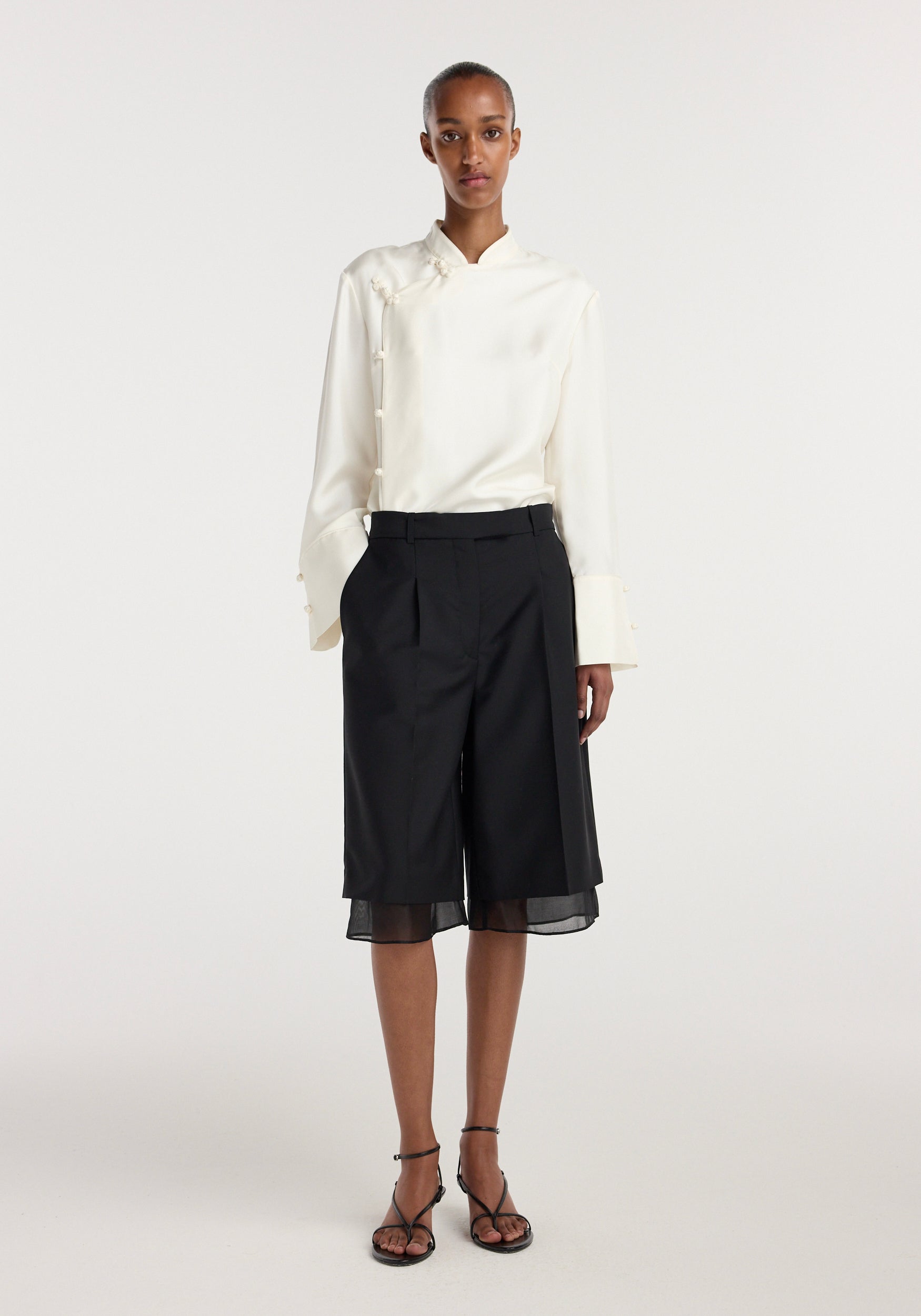 Model wearing elegant black Organza lined shorts | black paired with a long-sleeve white blouse and black strappy heels, standing against a plain white background.