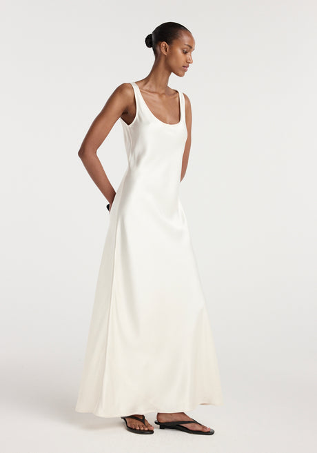 Model wearing a long, elegant scoop neck satin dress in ivory, holding a small black clutch and wearing black sandals, standing against a plain background.