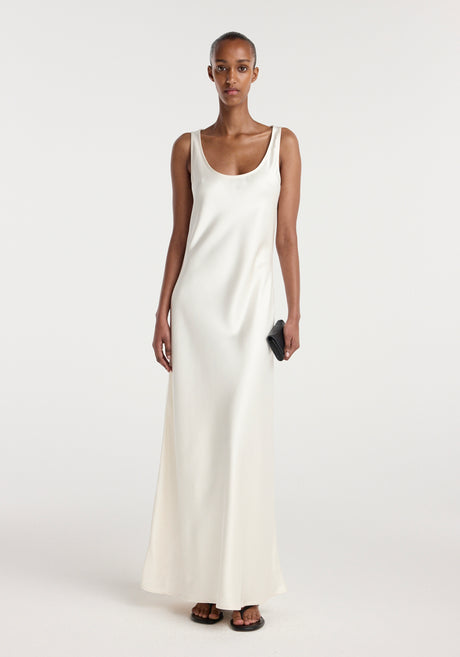 Model wearing a long, elegant scoop neck satin dress in ivory, holding a small black clutch and wearing black sandals, standing against a plain background.