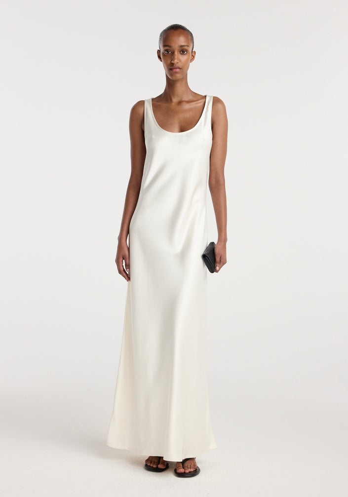 Model wearing a long, elegant scoop neck satin dress in ivory, holding a small black clutch and wearing black sandals, standing against a plain background.