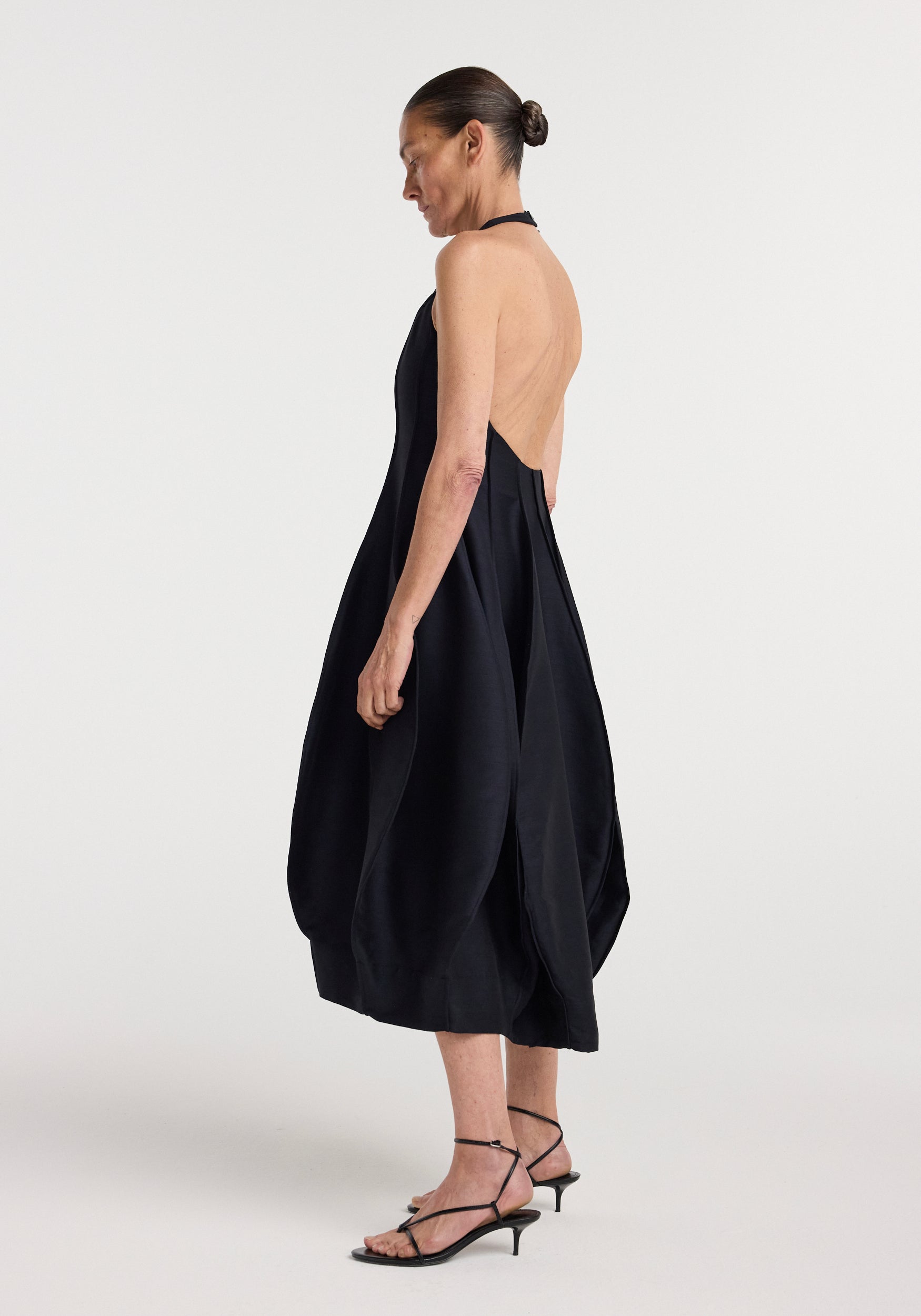 Woman wearing a black open back tulip dress | night with a halter neckline and balloon hem, paired with black strappy kitten heels, standing in profile against a plain background.