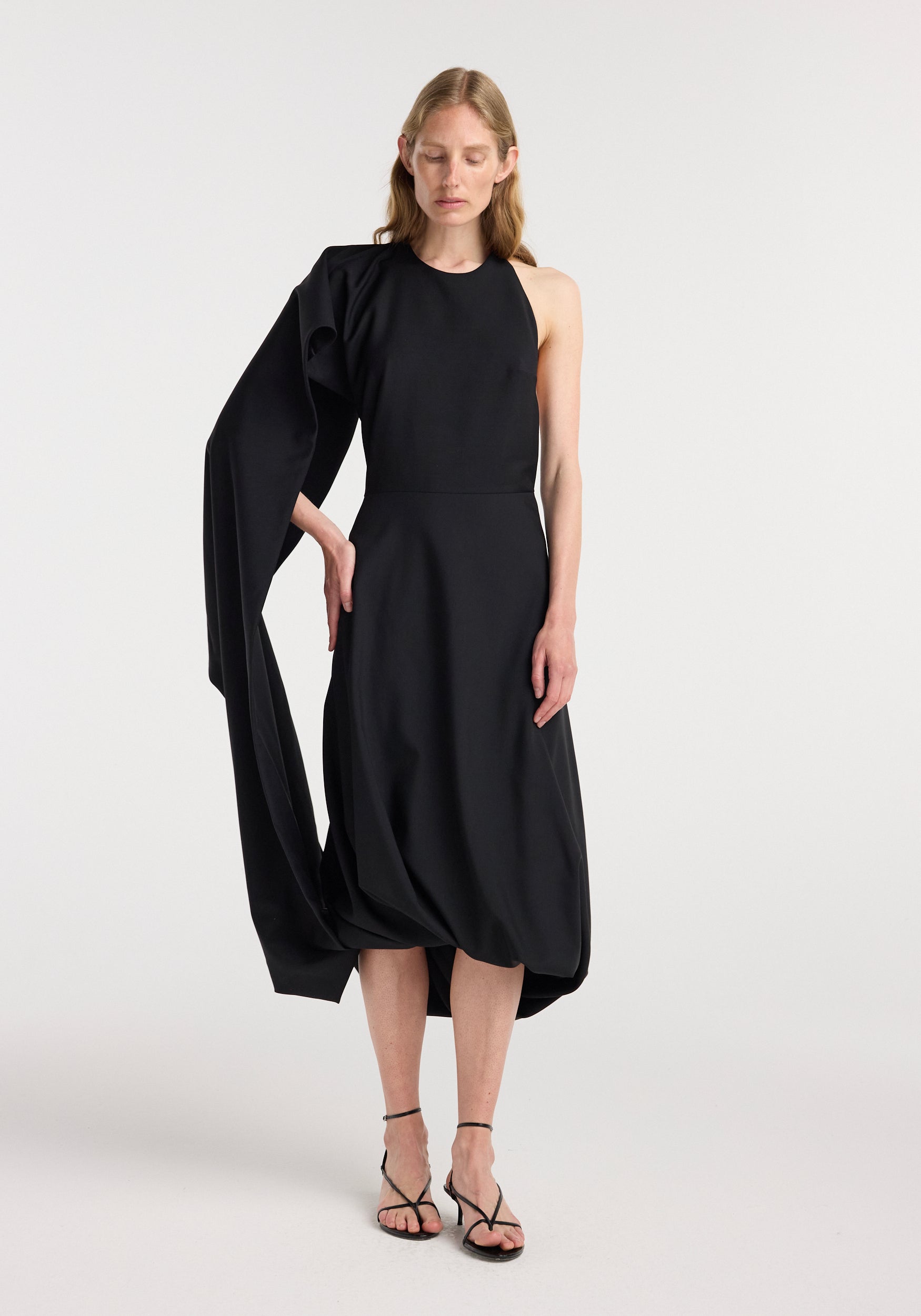 Model wearing a sleeveless black twisted cape dress | black with an asymmetrical flowing cape attached to one shoulder and strappy black high-heeled sandals.