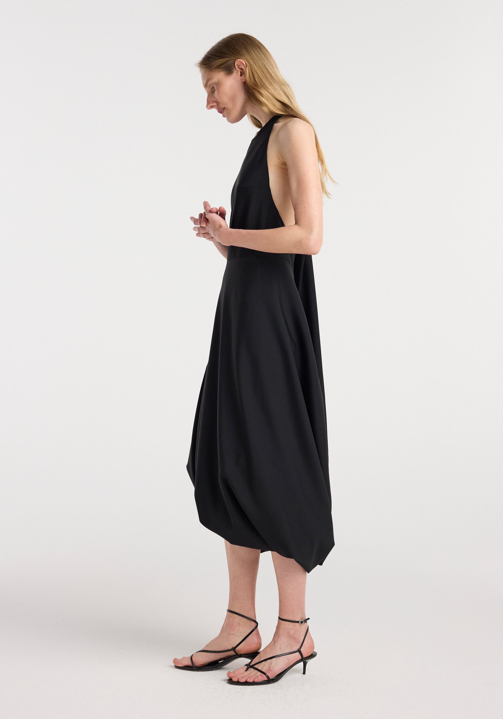 Woman wearing a sleeveless black twisted cape dress | black with a high neckline, side cutouts, and a draped asymmetrical hem paired with strappy black sandals.
