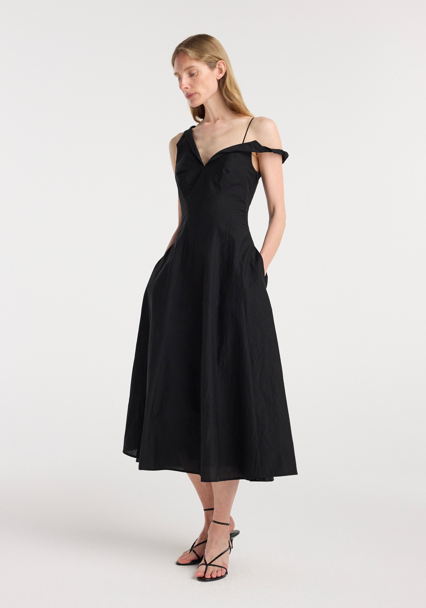 A woman wearing a black midi-length twisted shoulder dress | black with thin straps and an off-shoulder detail, styled with black strappy heels, standing against a plain white background.