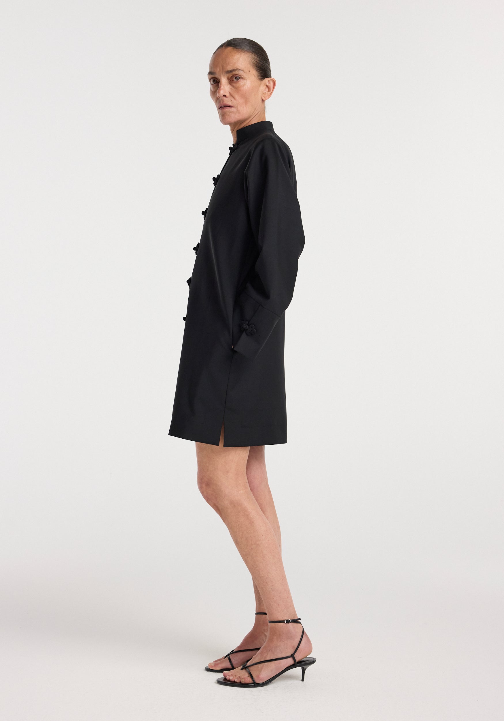 A woman wearing a black Pankou closure dress | black with long sleeves, a mandarin collar, and decorative button closures, paired with black strappy low-heeled sandals.