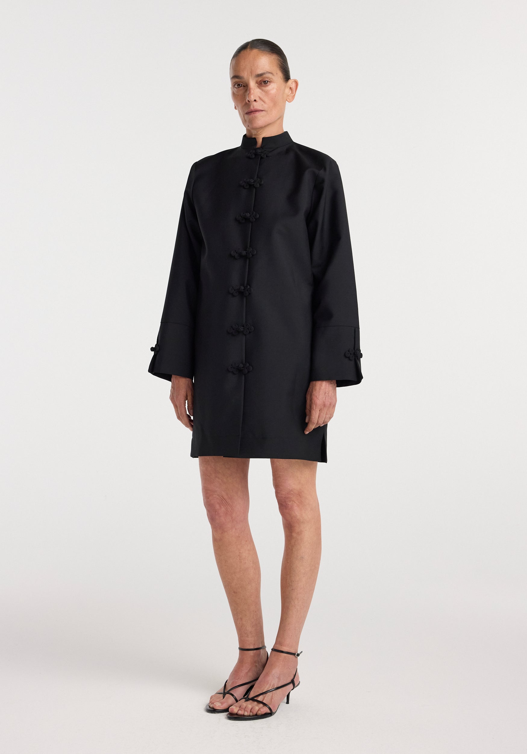 Woman wearing a black Pankou closure dress | black with long sleeves, standing against a plain white background, paired with black strappy heels.