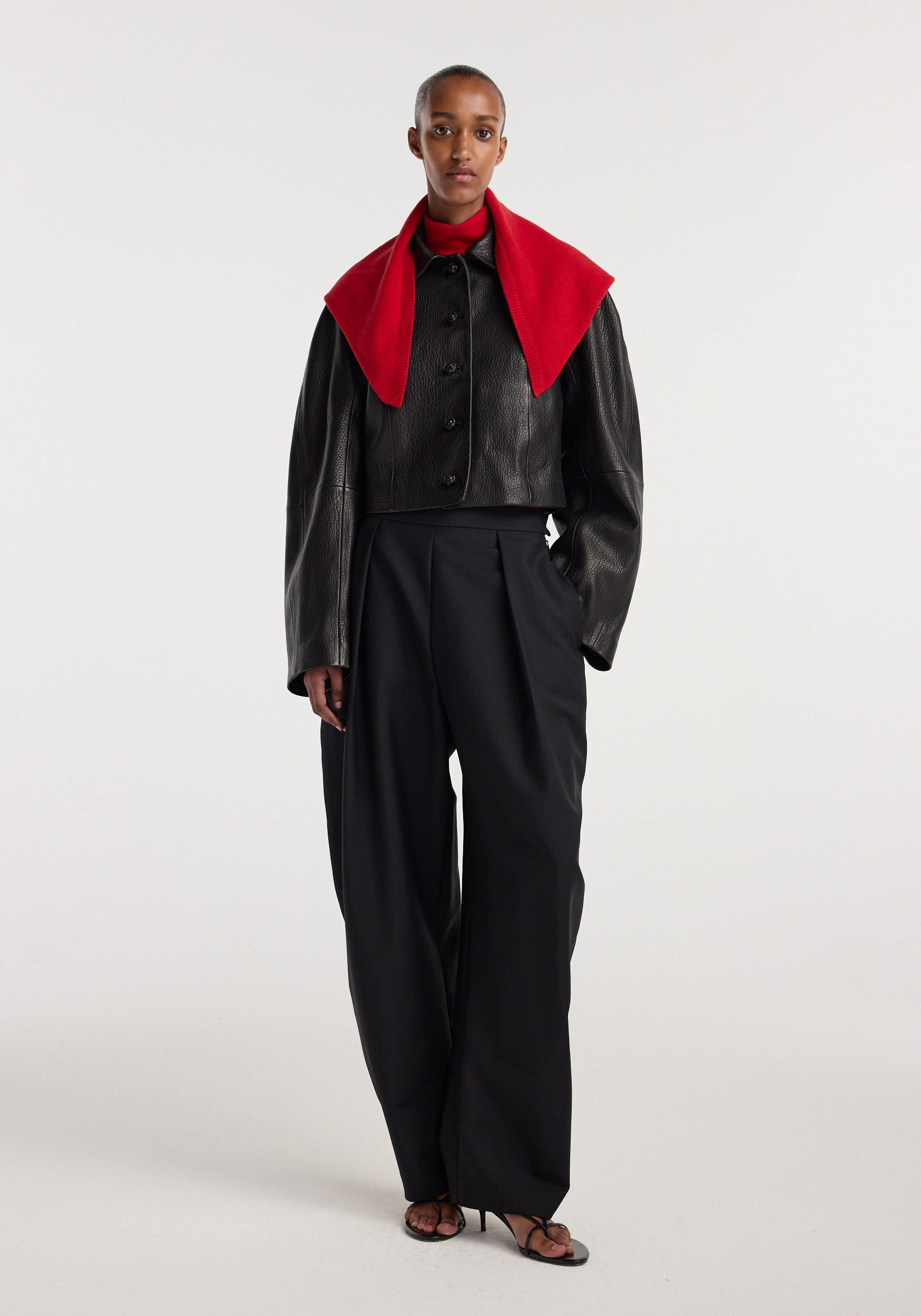 Model wearing a small knitted triangle scarf in red over a black leather cropped jacket and high-waisted black wide-leg trousers with heeled sandals.