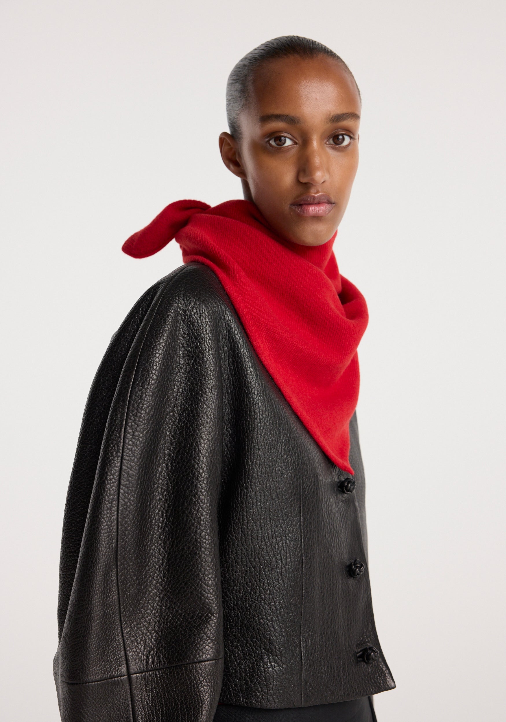 Model wearing a vibrant red small knitted triangle scarf | red tied loosely around the neck, paired with a textured black leather jacket against a plain background.