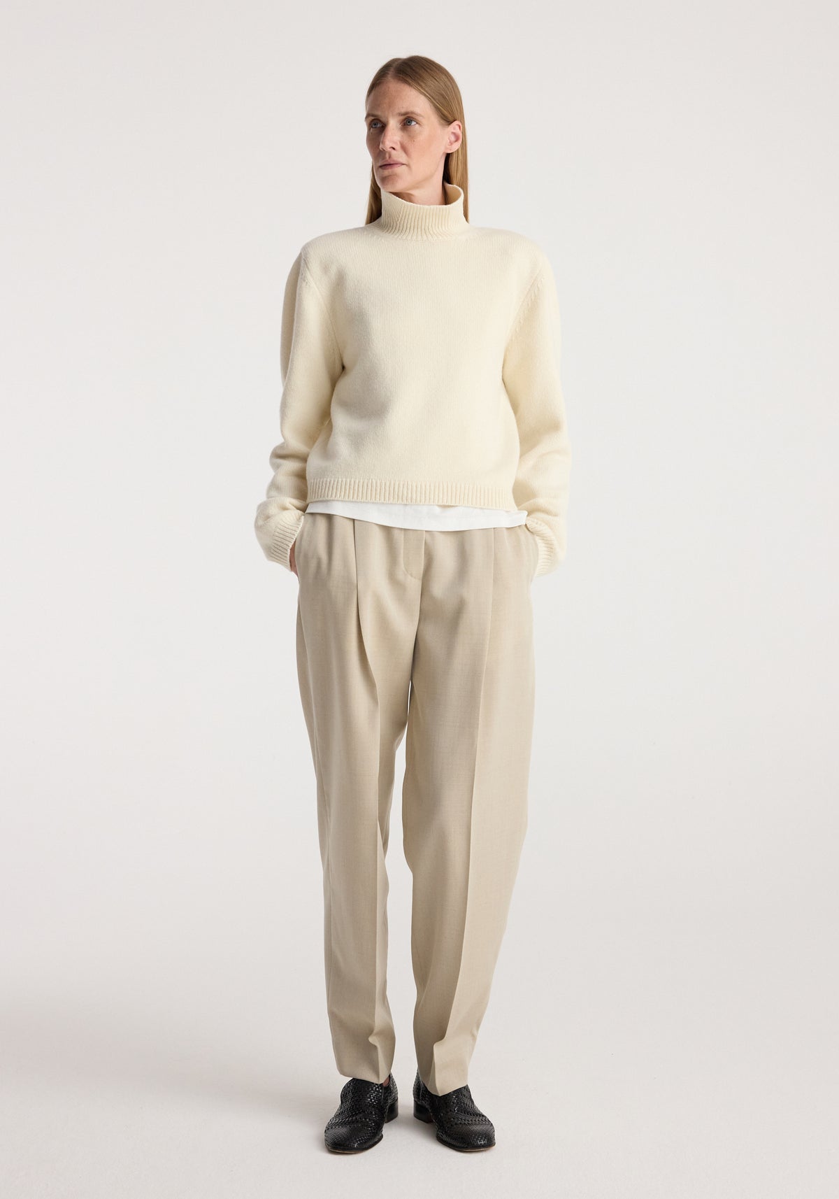 Woman wearing a cream Statement shoulder cropped turtleneck with long sleeves, paired with beige pleated trousers and black shoes, standing against a plain background.