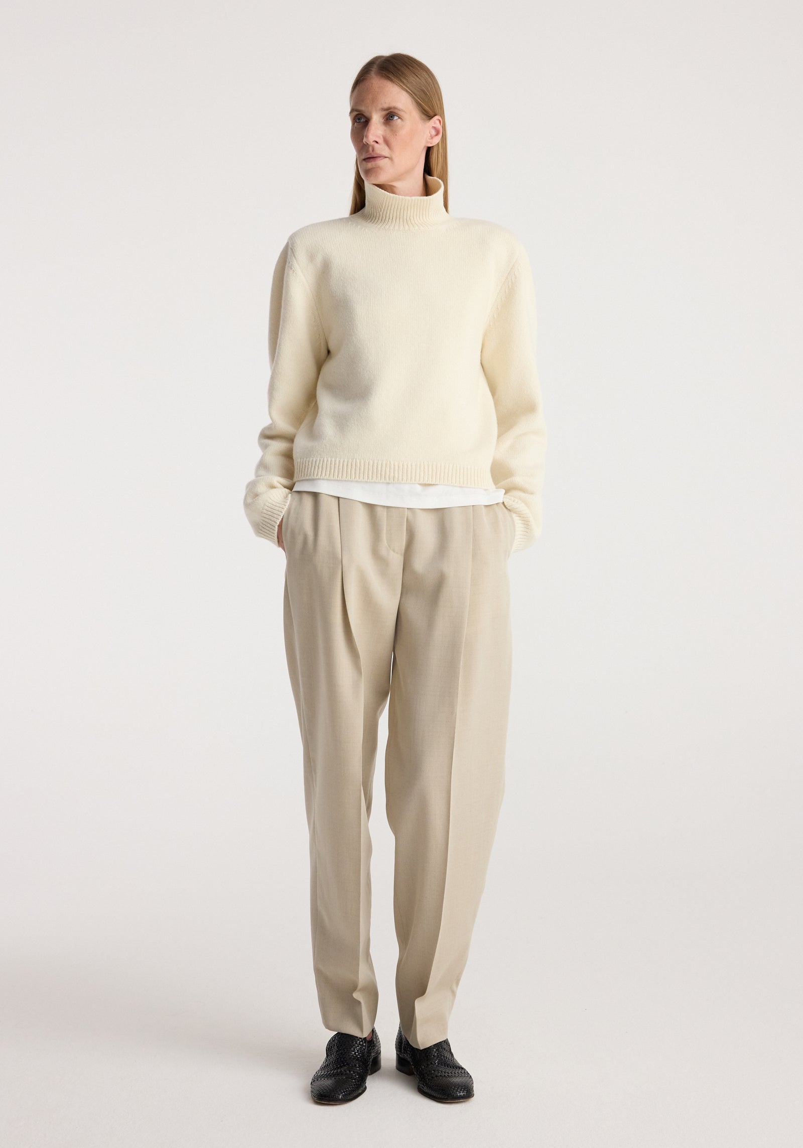 Woman wearing a cream Statement shoulder cropped turtleneck with long sleeves, paired with beige pleated trousers and black shoes, standing against a plain background.