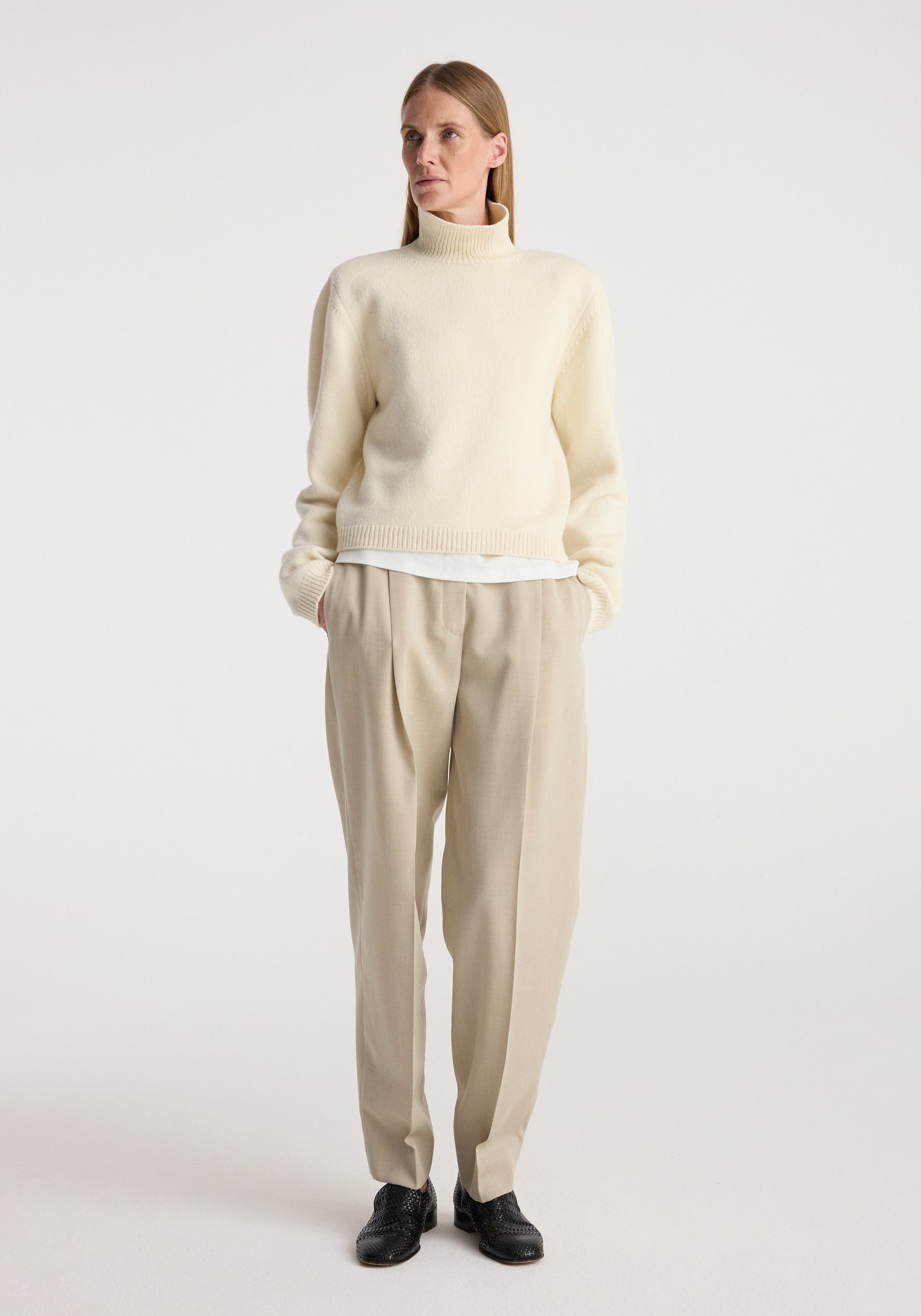 Woman wearing a cream Statement shoulder cropped turtleneck with long sleeves, paired with beige pleated trousers and black shoes, standing against a plain background.