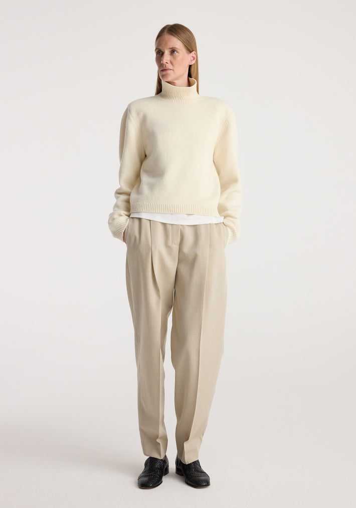 Woman wearing a cream Statement shoulder cropped turtleneck with long sleeves, paired with beige pleated trousers and black shoes, standing against a plain background.