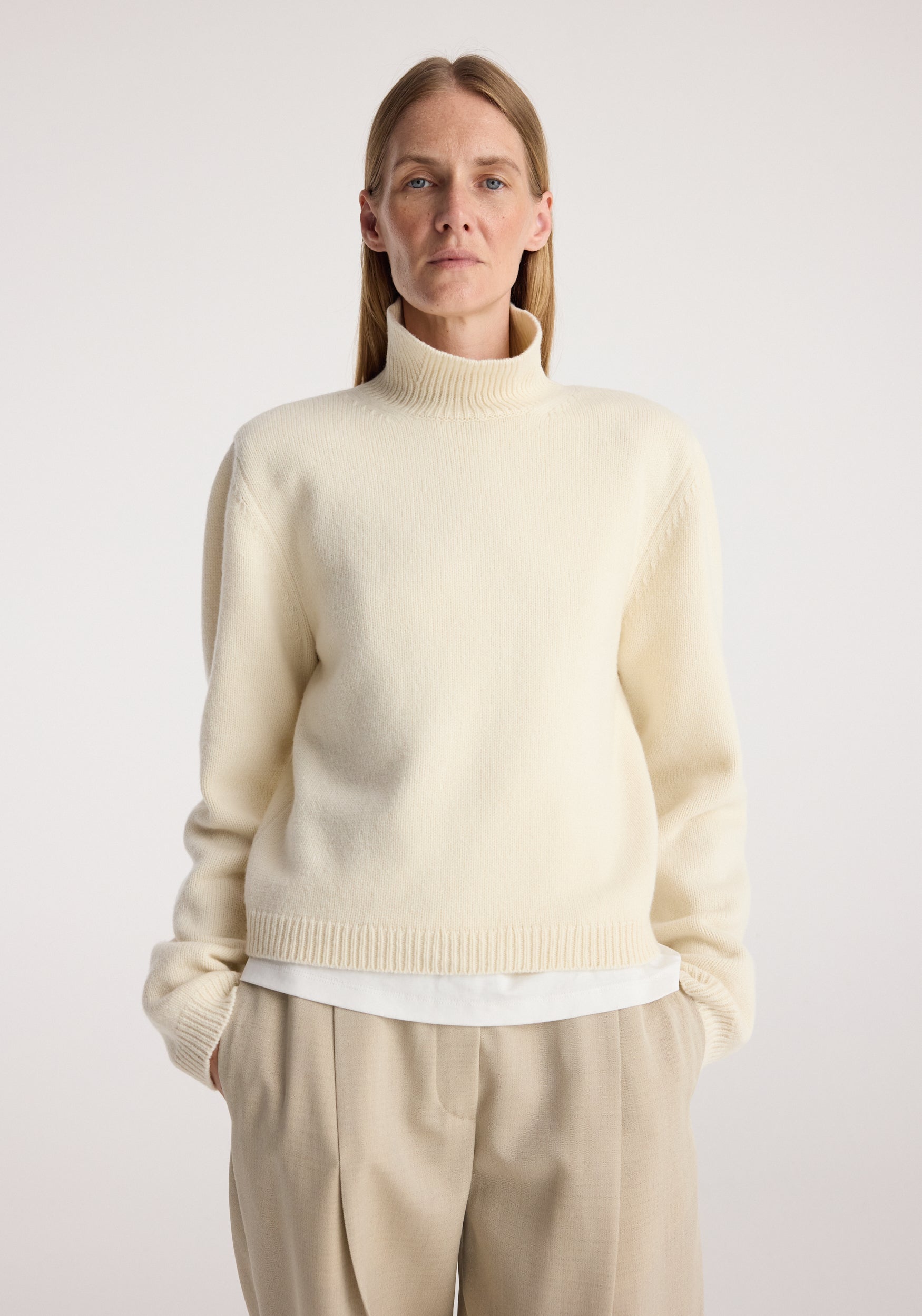 Woman wearing a cream Statement shoulder cropped turtleneck | cream with long sleeves and a high neckline, paired with beige trousers against a plain background.