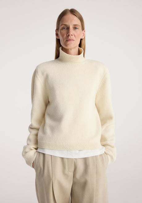 Woman wearing a cream Statement shoulder cropped turtleneck with long sleeves, paired with beige pleated trousers and black shoes, standing against a plain background.