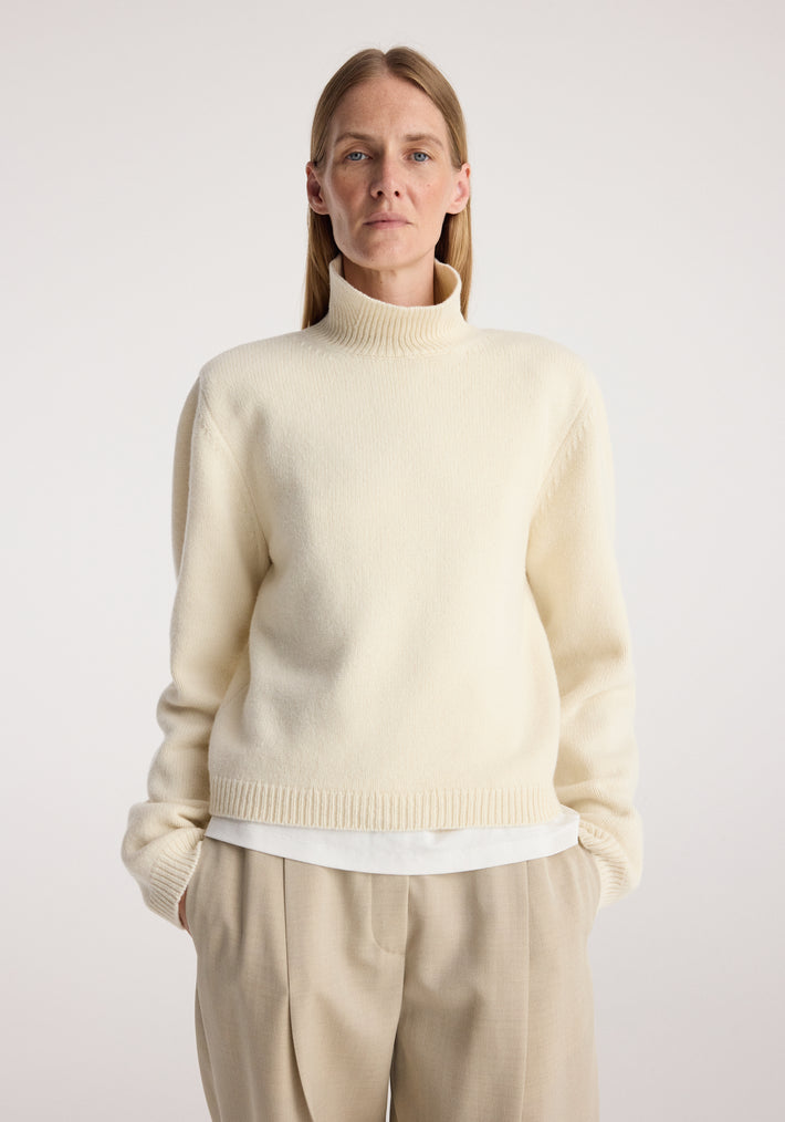 Woman wearing a cream Statement shoulder cropped turtleneck | cream with long sleeves and a high neckline, paired with beige trousers against a plain background.