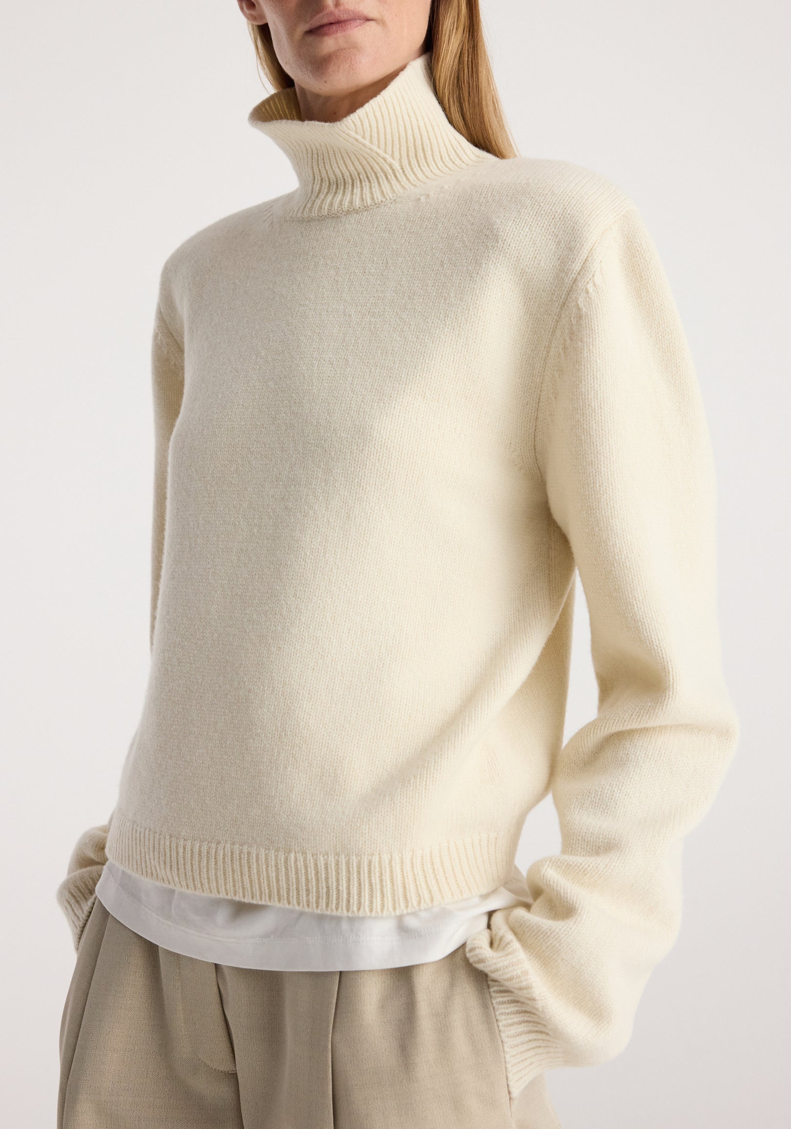 Woman wearing a cream-colored Statement shoulder cropped turtleneck | cream with long sleeves and ribbed cuffs, paired with beige trousers and a white underlayer shirt.
