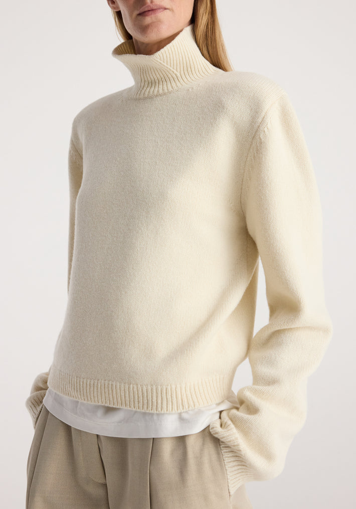 Woman wearing a cream-colored Statement shoulder cropped turtleneck | cream with long sleeves and ribbed cuffs, paired with beige trousers and a white underlayer shirt.
