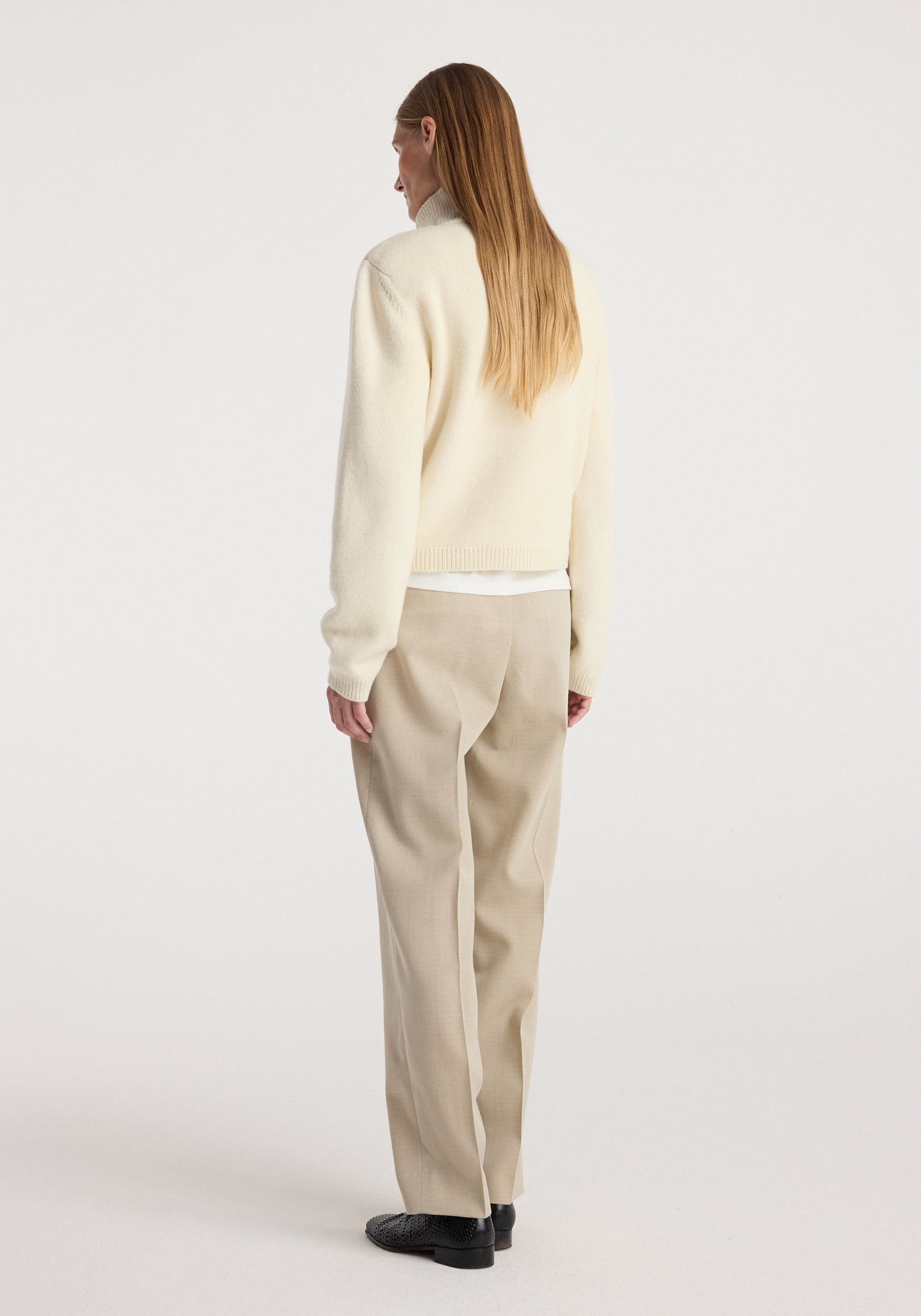 Woman wearing a cream statement shoulder cropped turtleneck with long sleeves, paired with beige trousers and black shoes, standing facing away against a white background.