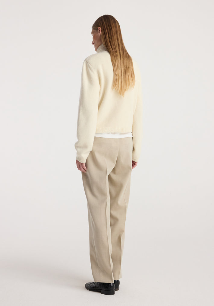 Woman wearing a cream statement shoulder cropped turtleneck with long sleeves, paired with beige trousers and black shoes, standing facing away against a white background.
