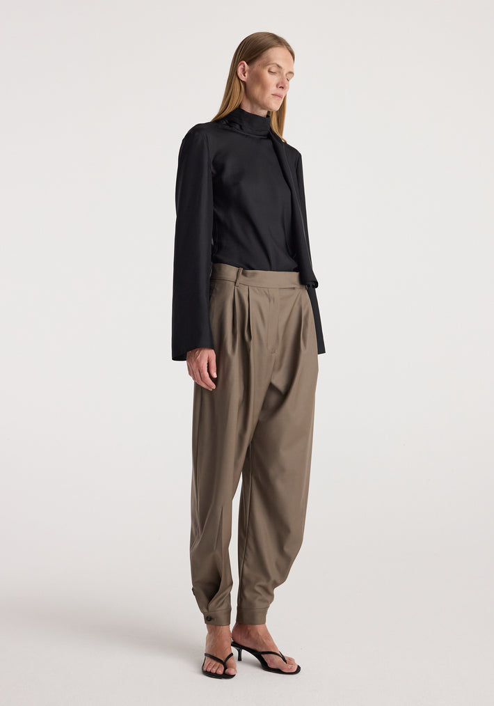 Woman wearing tailored cuff trousers | desert brown paired with a black high-neck top and black open-toe heels against a plain light background.