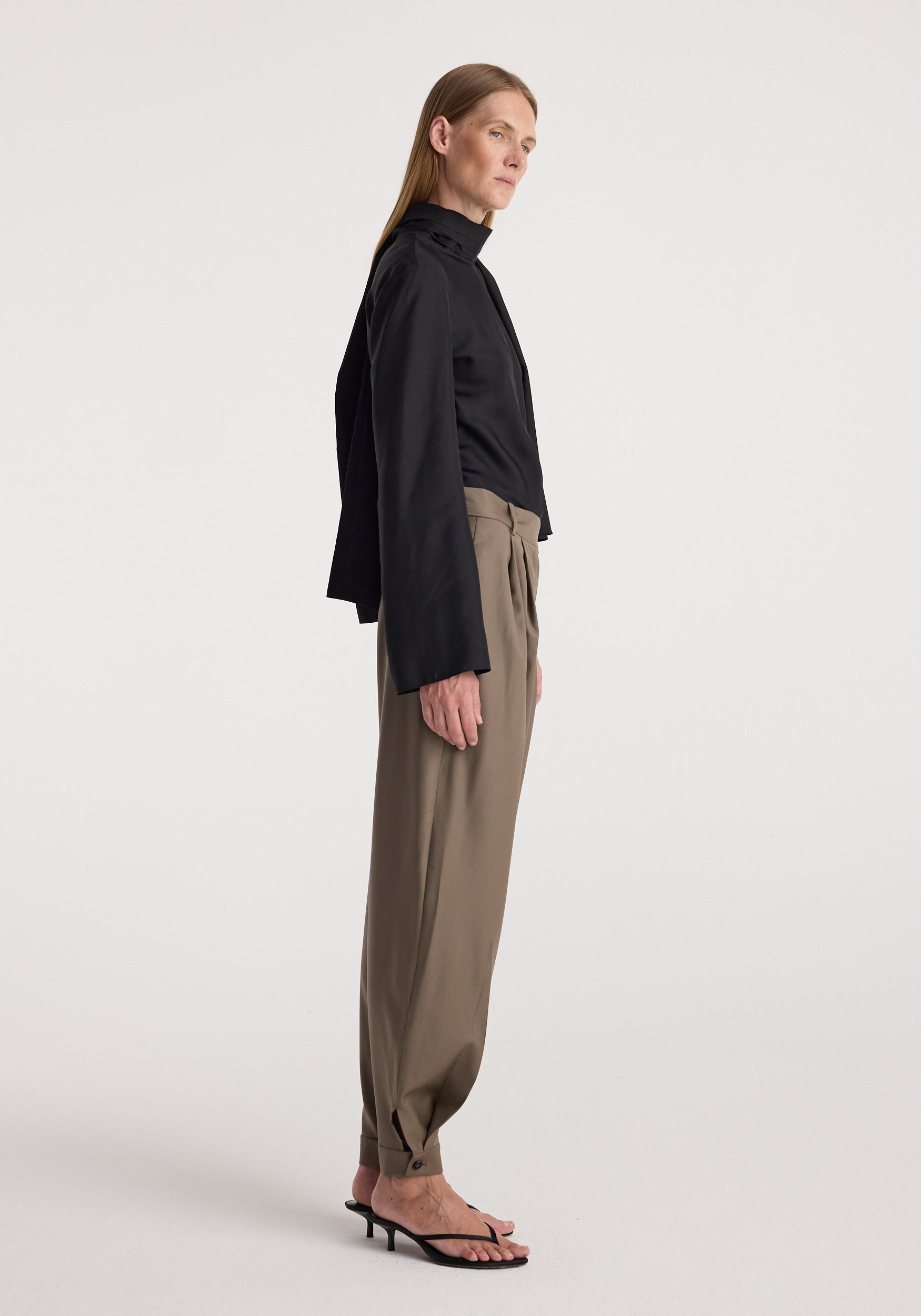 Woman standing in profile wearing tailored cuff trousers | desert brown paired with a loose black long-sleeve top and black heeled sandals against a plain background.