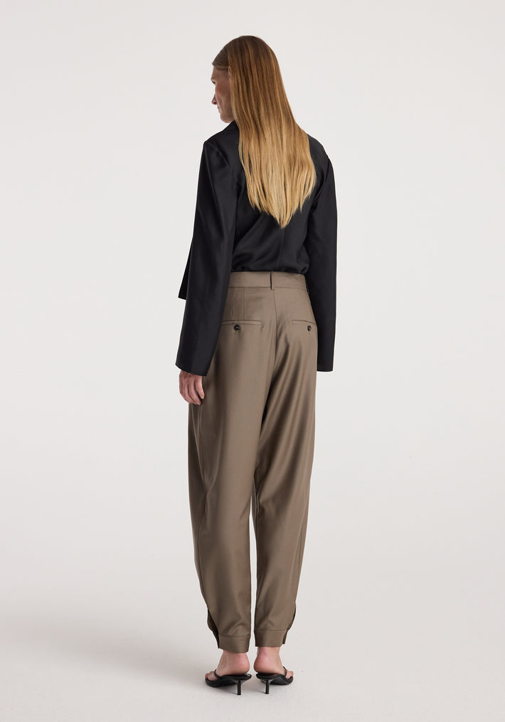 Woman wearing tailored cuff trousers | desert brown paired with a black long-sleeve top and black open-heeled shoes, standing with her back to the camera.