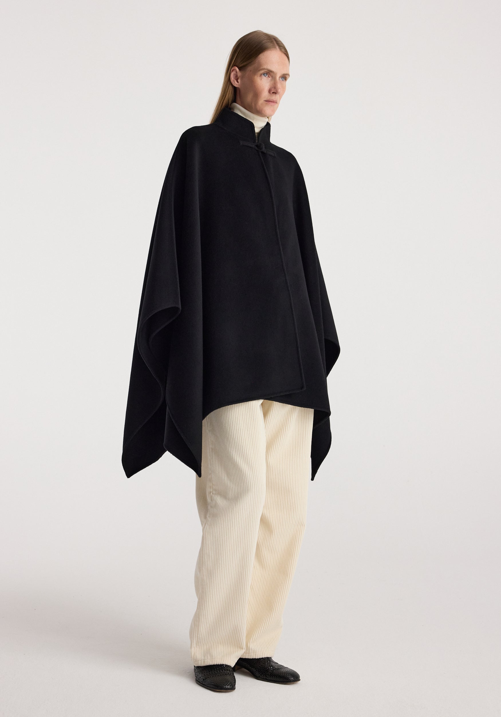 Woman wearing a Double-faced pankou poncho | black over a white turtleneck and cream pants, standing against a plain light background.