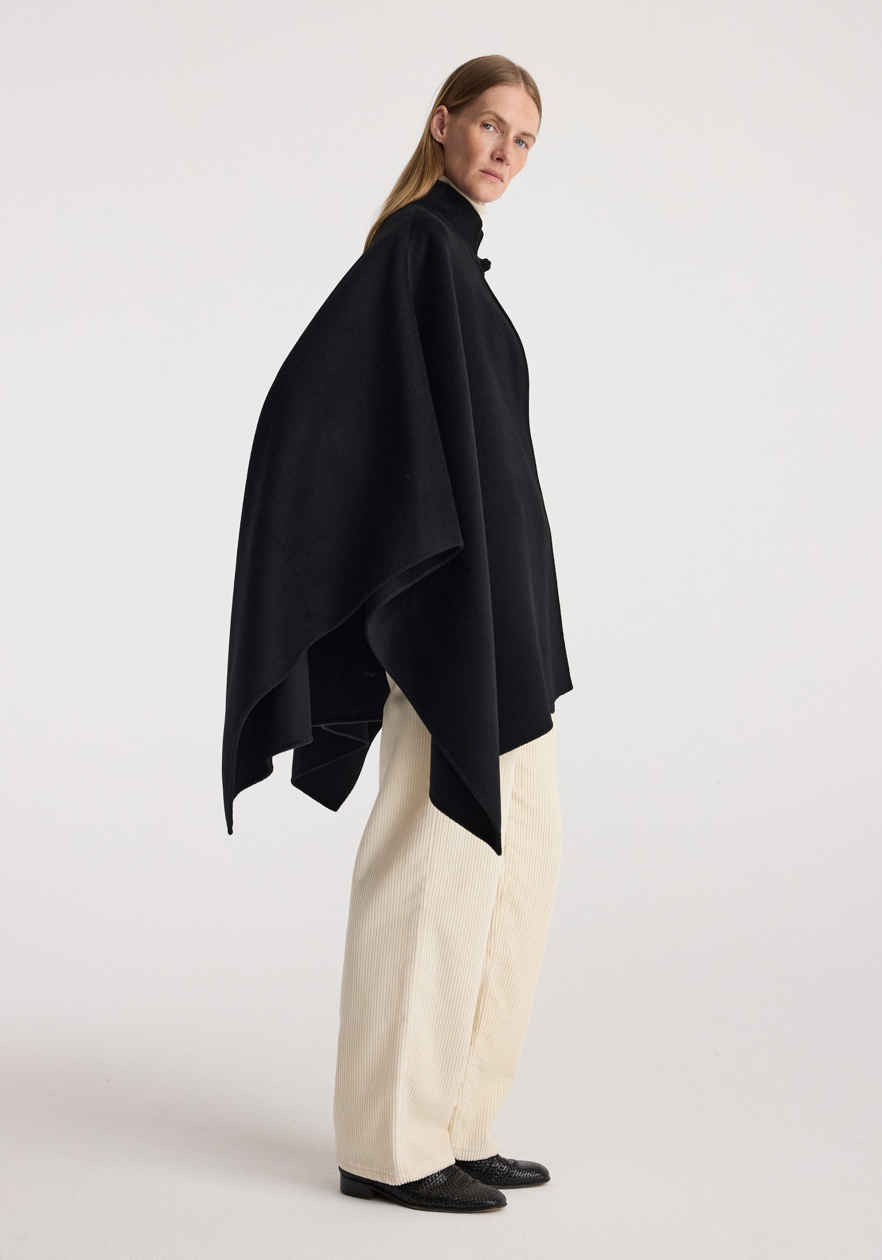 Woman wearing a black Double-faced pankou poncho | black, standing in a side profile against a plain background, paired with cream-colored pants and black shoes.