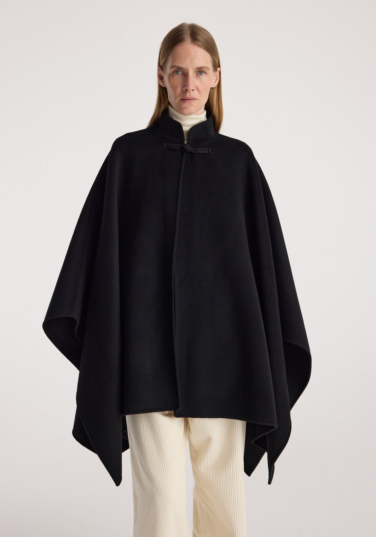 Woman wearing a black double-faced pankou poncho | black over a white turtleneck and cream pants, standing against a plain light background.