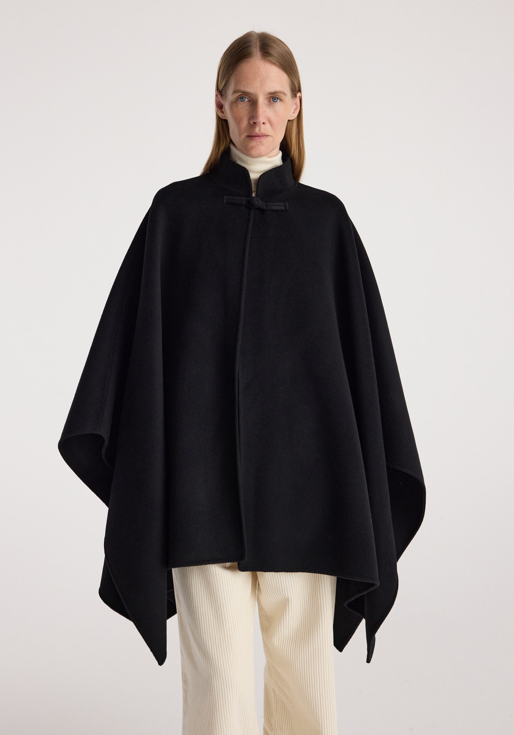Woman wearing a black double-faced pankou poncho | black over a white turtleneck and cream pants, standing against a plain light background.