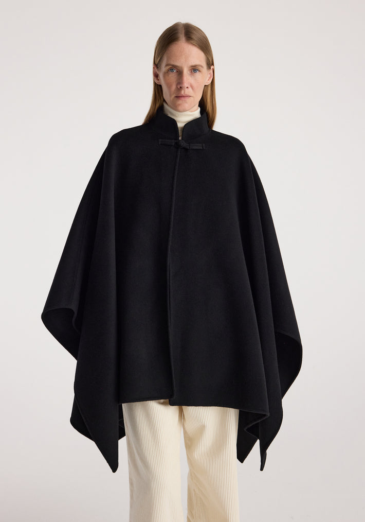Woman wearing a black double-faced pankou poncho | black over a white turtleneck and cream pants, standing against a plain light background.