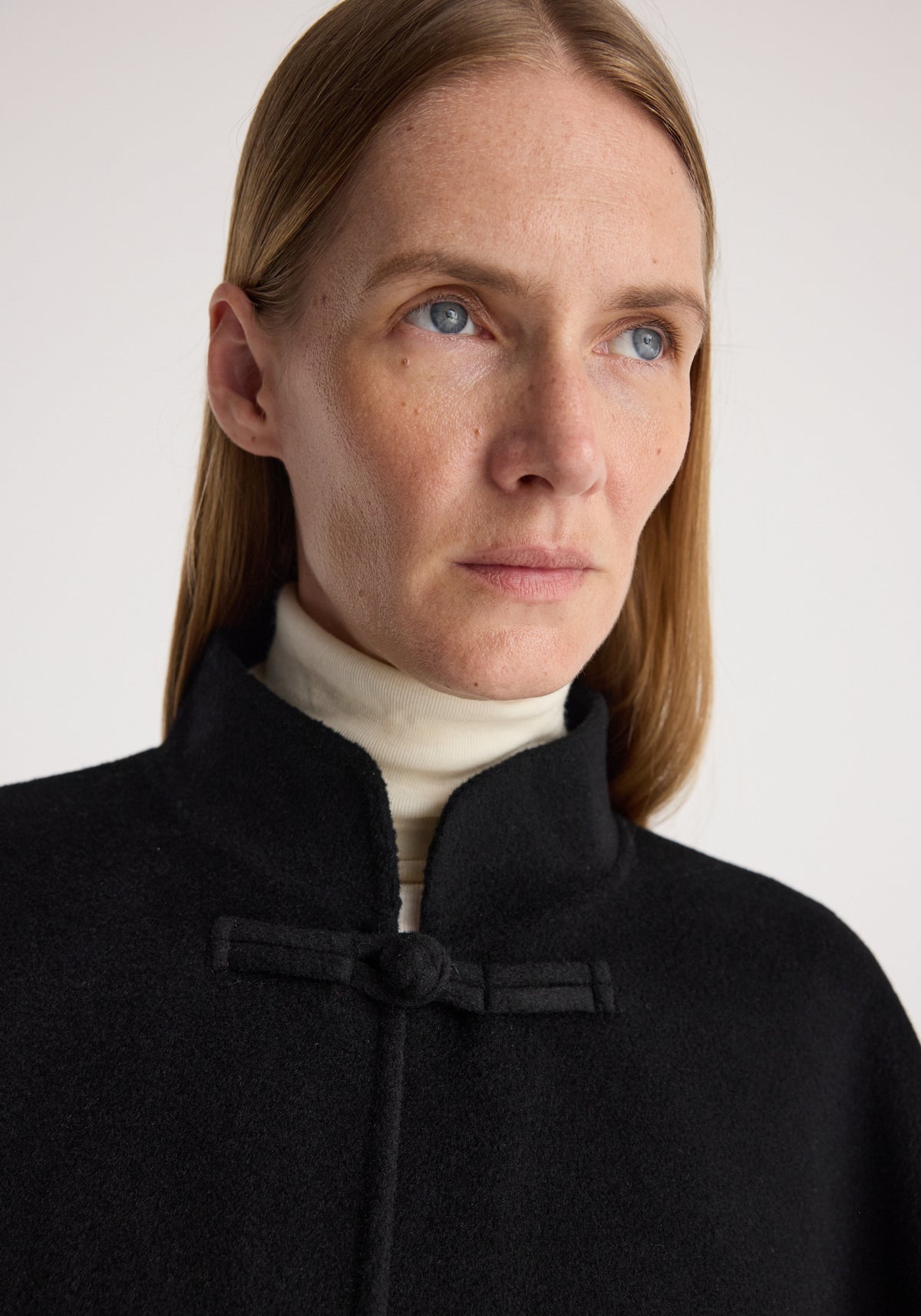 Close-up of a woman with light skin and blue eyes wearing a Double-faced pankou poncho | black over a white turtleneck, showing the high collar and pankou knot detail.
