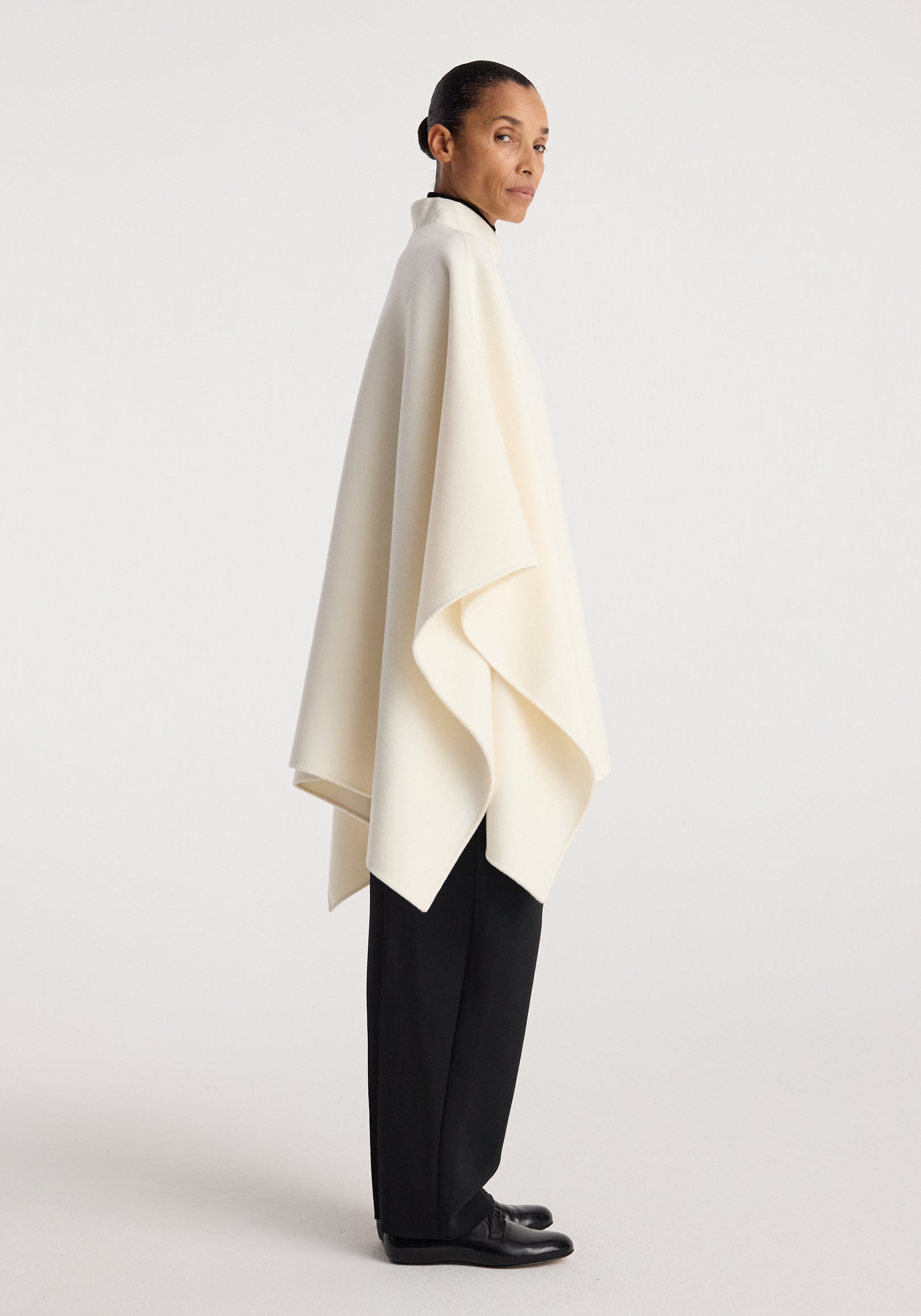 Side view of a model wearing a double-faced pankou poncho | off white, draped loosely over black pants and black shoes against a plain background.