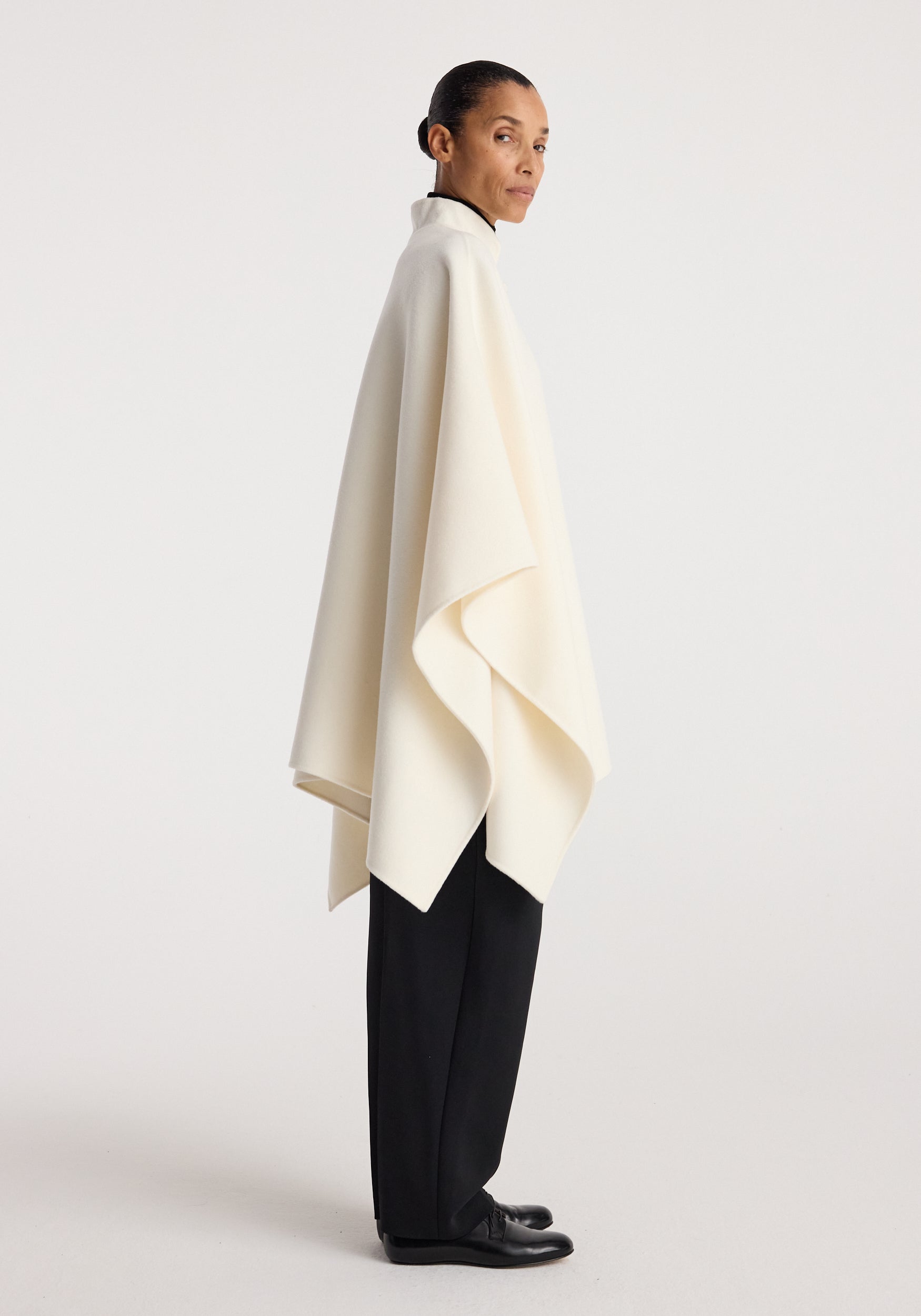 Side view of a model wearing a double-faced pankou poncho | off white, draped loosely over black pants and black shoes against a plain background.