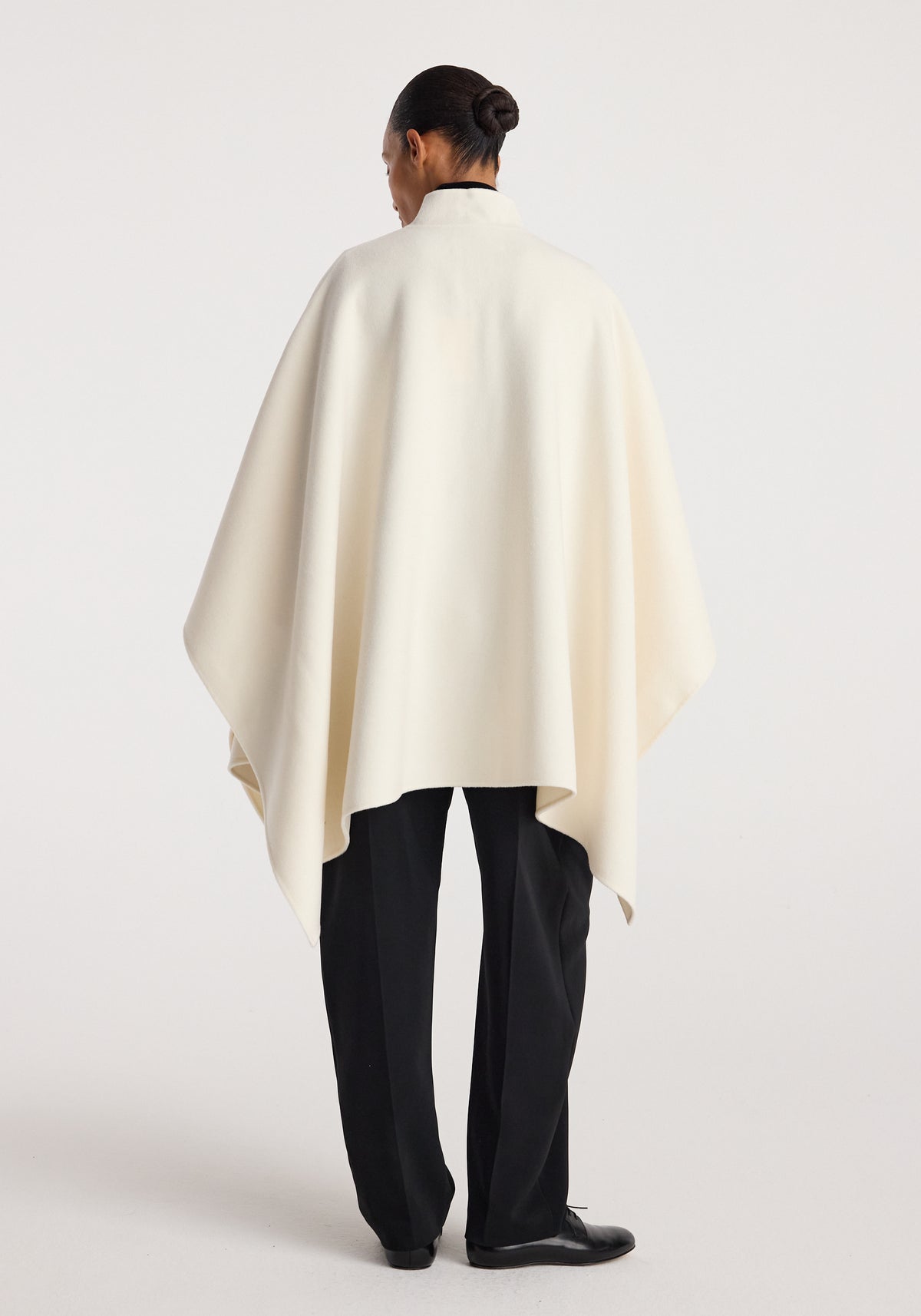 Model standing with back facing the camera wearing a Double-faced pankou poncho | off white over black trousers and black shoes against a plain background.