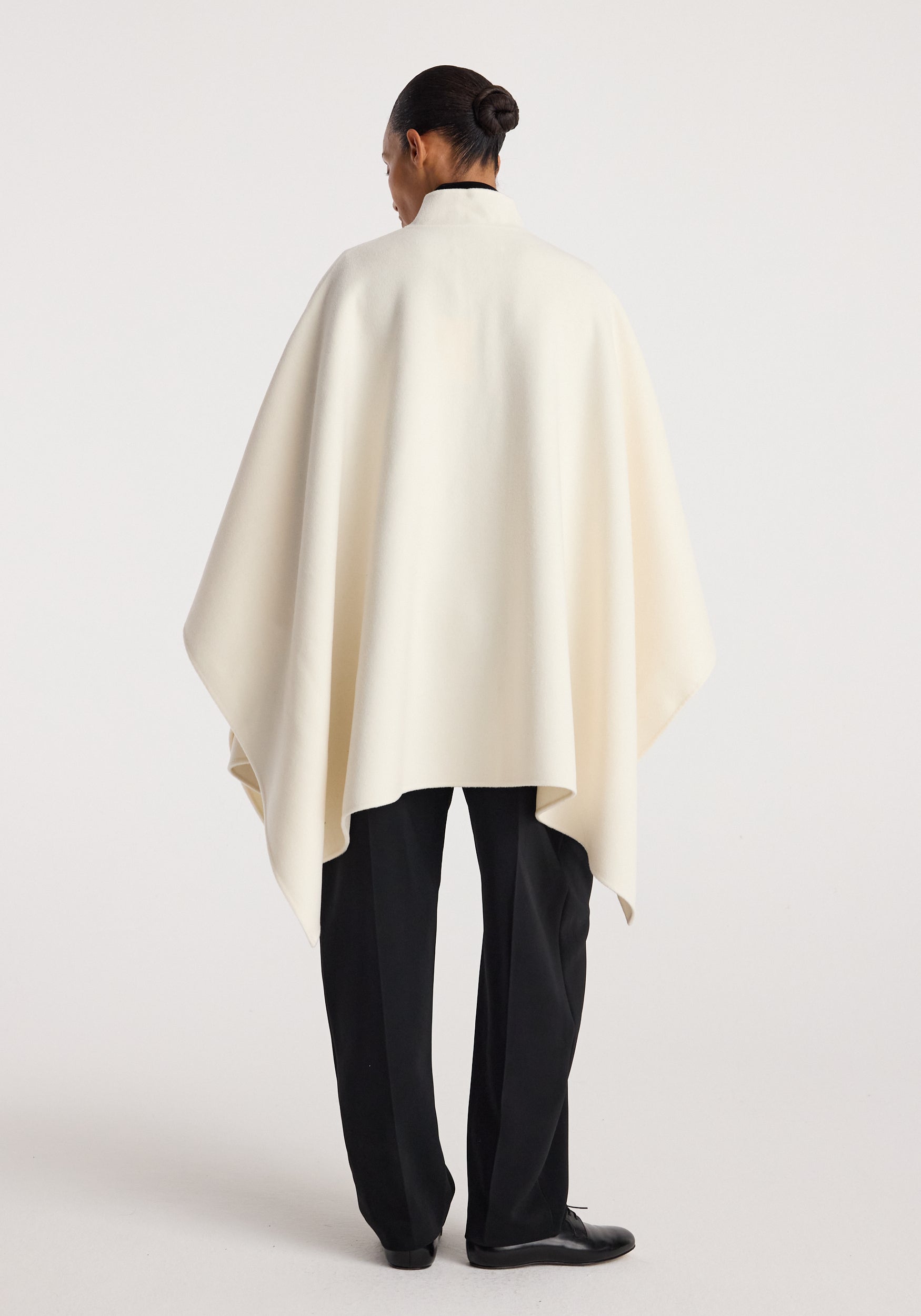 Model standing with back facing the camera wearing a Double-faced pankou poncho | off white over black trousers and black shoes against a plain background.