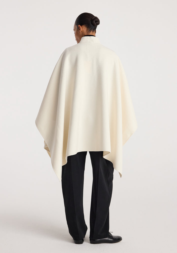 Model standing with back facing the camera wearing a Double-faced pankou poncho | off white over black trousers and black shoes against a plain background.