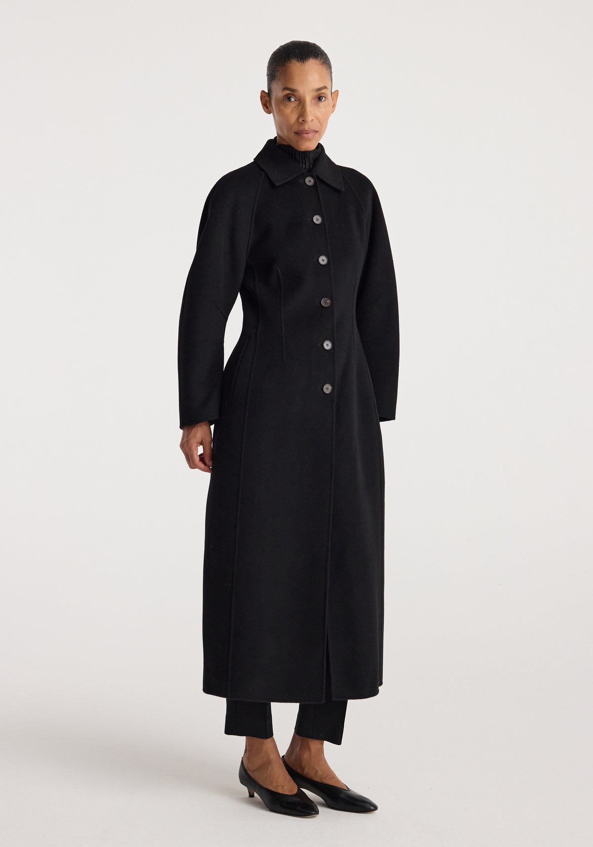 Woman wearing a long black buttoned coat with a structured silhouette and collar, standing against a plain white background, showcasing the Double-faced elongated sculptural coat | black.