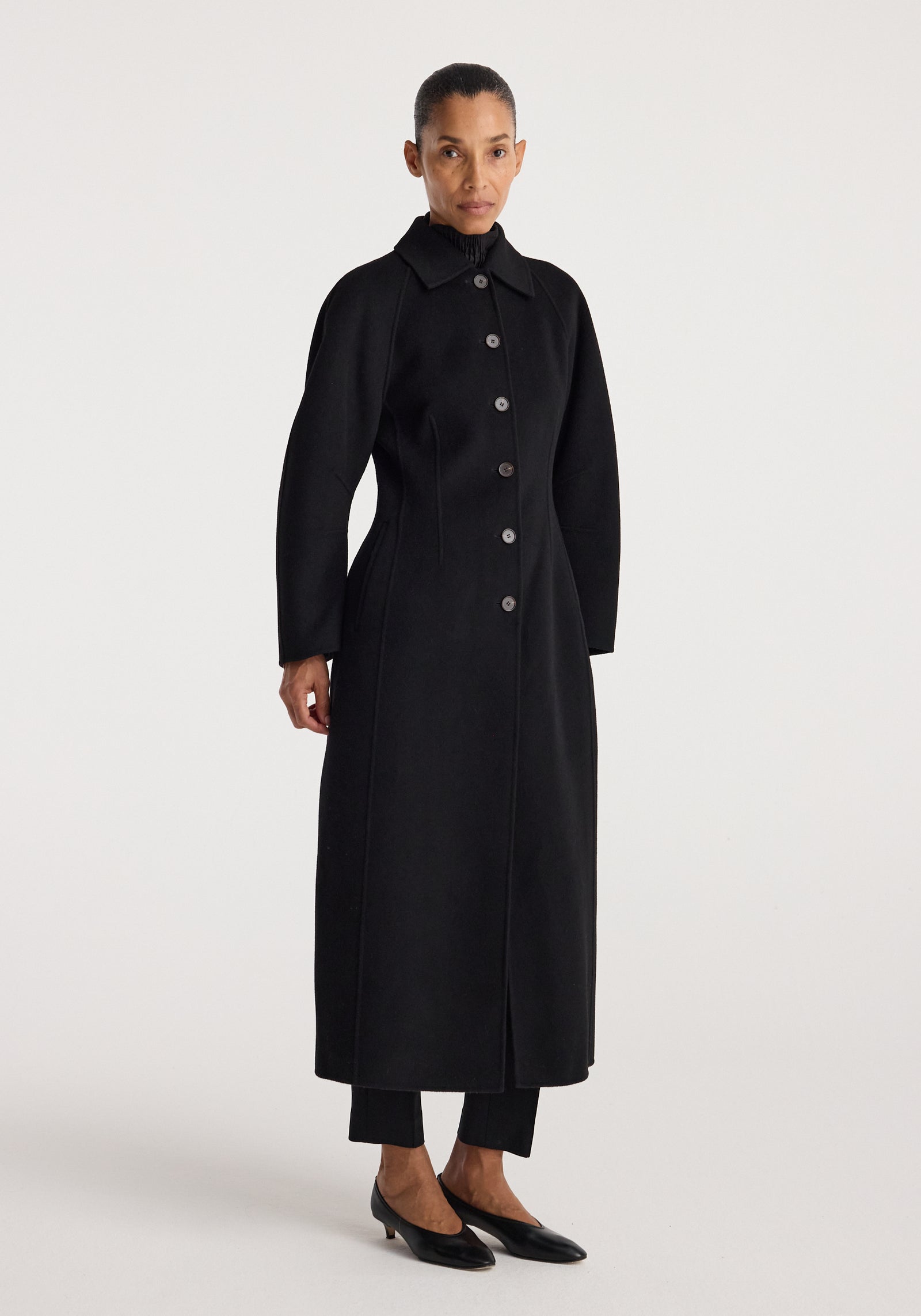 Woman wearing a long black buttoned coat with a structured silhouette and collar, standing against a plain white background, showcasing the Double-faced elongated sculptural coat | black.
