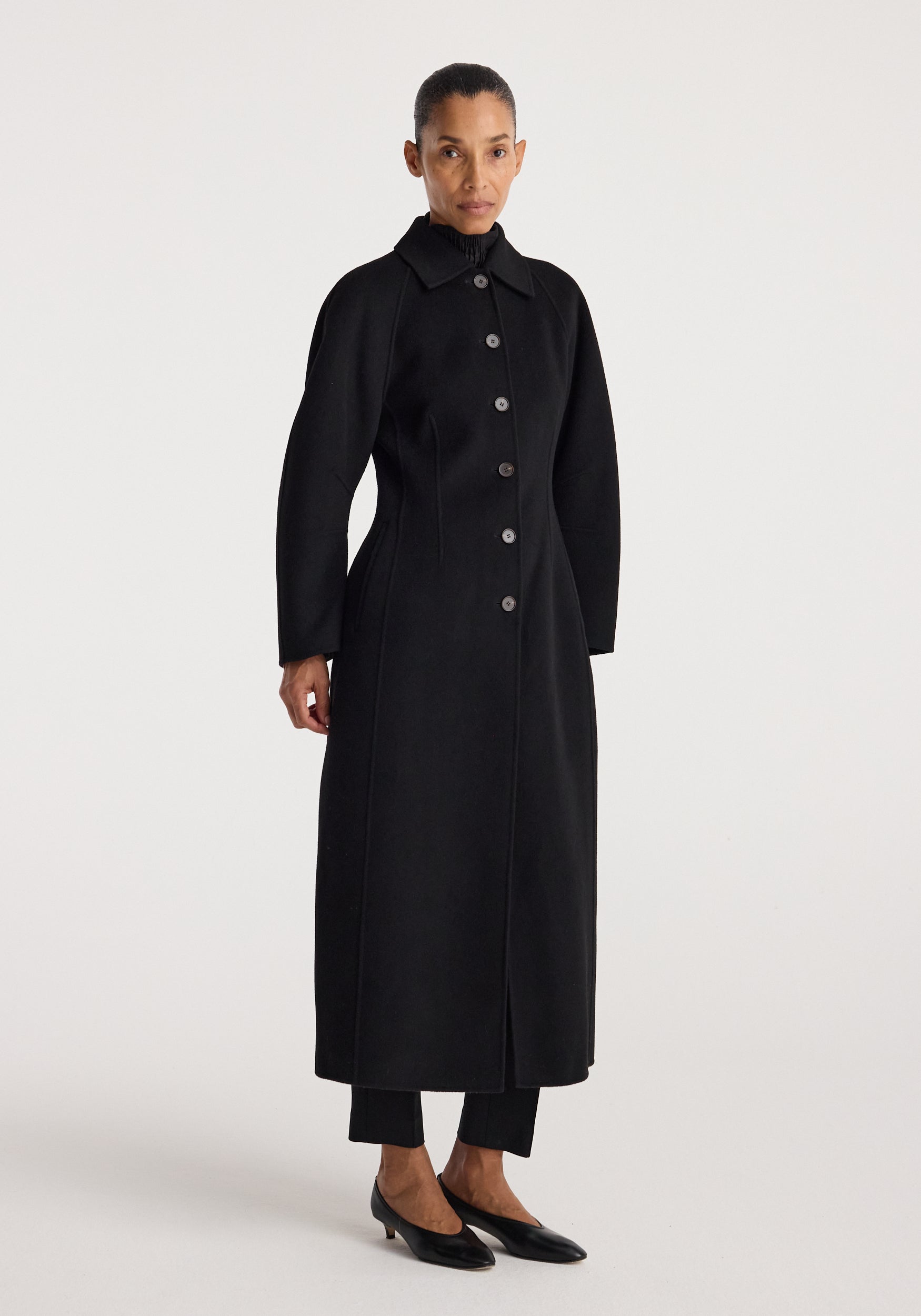 Woman wearing a long black buttoned coat with a structured silhouette and collar, standing against a plain white background, showcasing the Double-faced elongated sculptural coat | black.