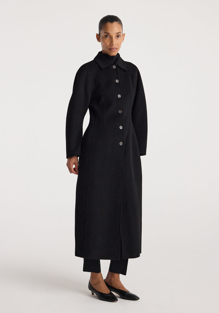 Woman wearing a long black buttoned coat with a structured silhouette and collar, standing against a plain white background, showcasing the Double-faced elongated sculptural coat | black.