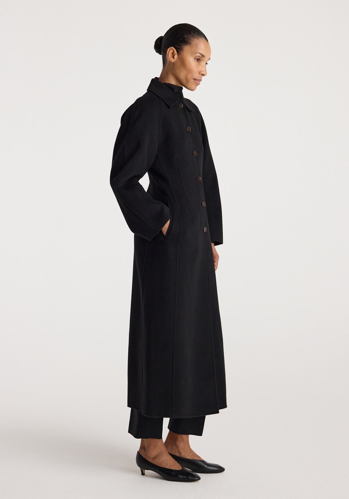 Woman wearing a minimalist, long black coat with buttons and side pockets, styled with black pants and low black heels, showcasing the Double-faced elongated sculptural coat | black.