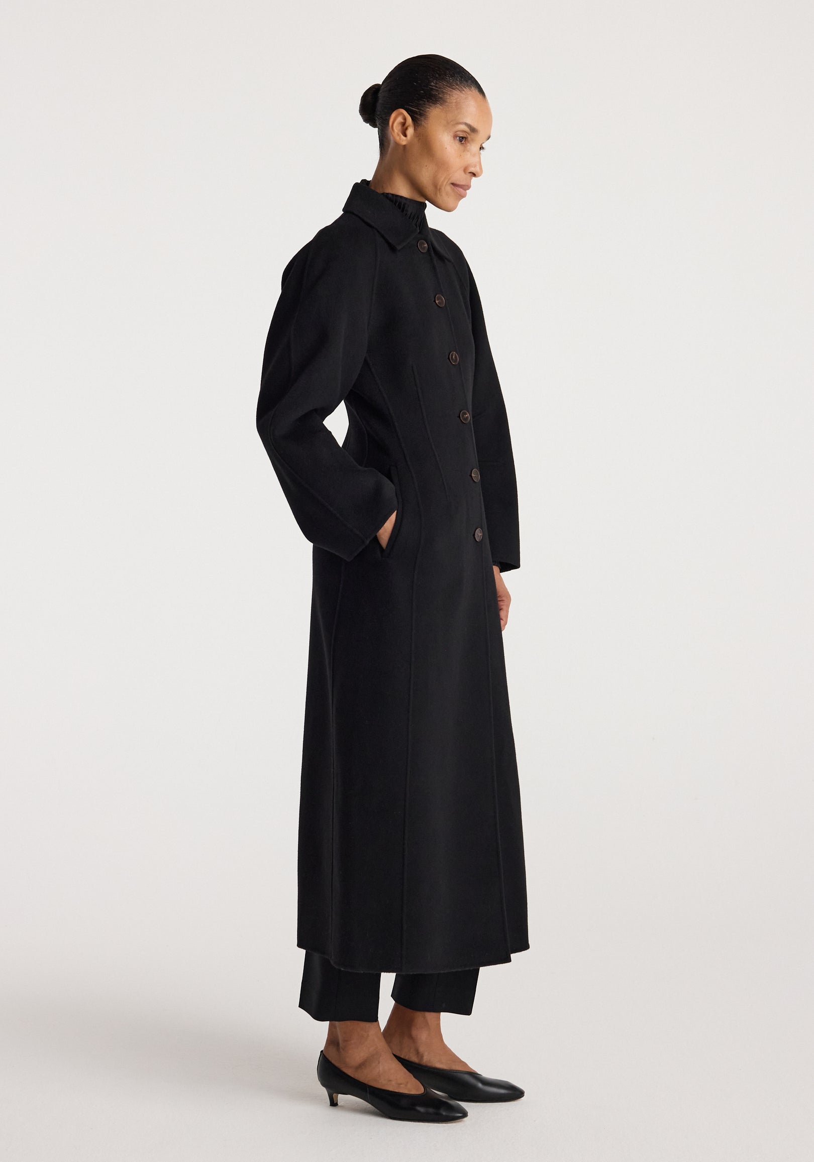 Woman wearing a minimalist, long black coat with buttons and side pockets, styled with black pants and low black heels, showcasing the Double-faced elongated sculptural coat | black.