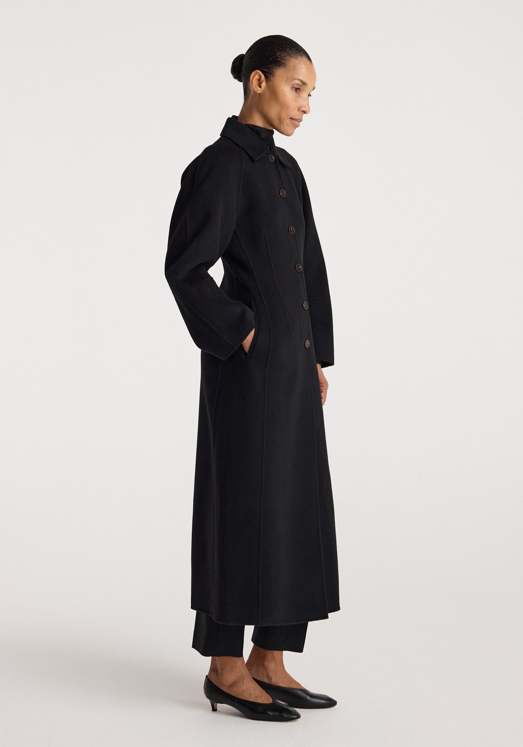 Woman wearing a minimalist, long black coat with buttons and side pockets, styled with black pants and low black heels, showcasing the Double-faced elongated sculptural coat | black.