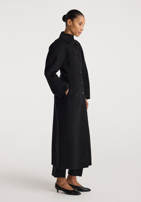 Woman wearing a long black buttoned coat with a structured silhouette and collar, standing against a plain white background, showcasing the Double-faced elongated sculptural coat | black.