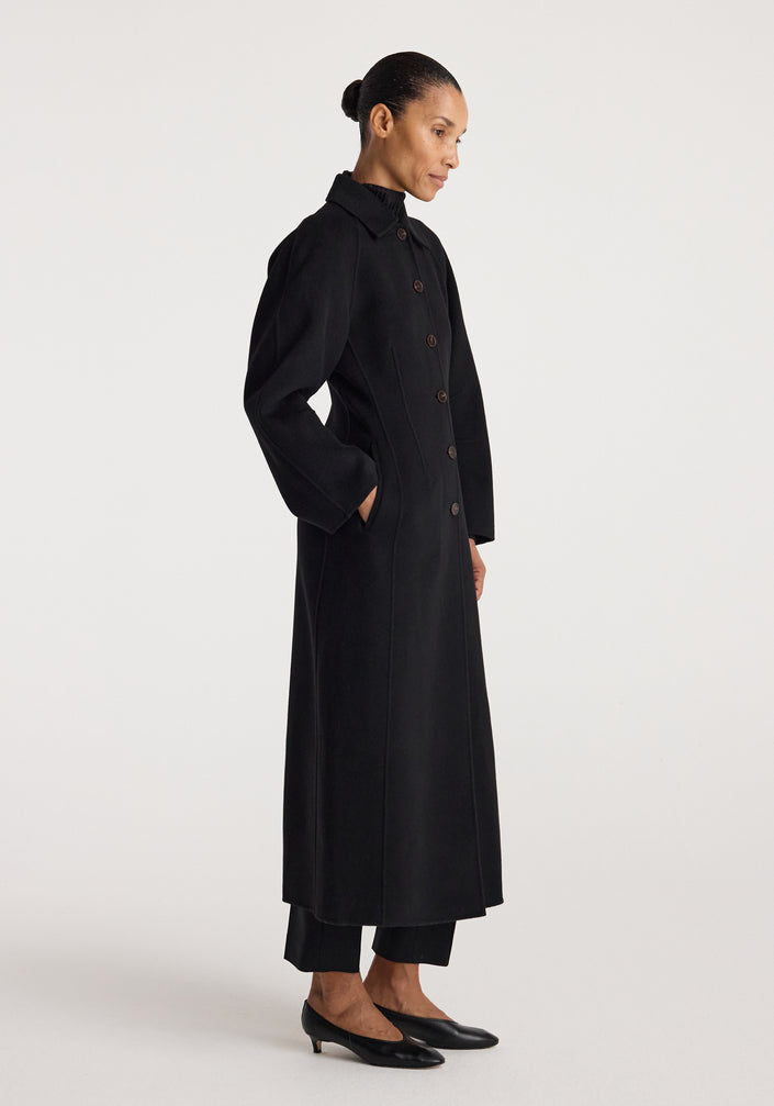 Woman wearing a long black buttoned coat with a structured silhouette and collar, standing against a plain white background, showcasing the Double-faced elongated sculptural coat | black.