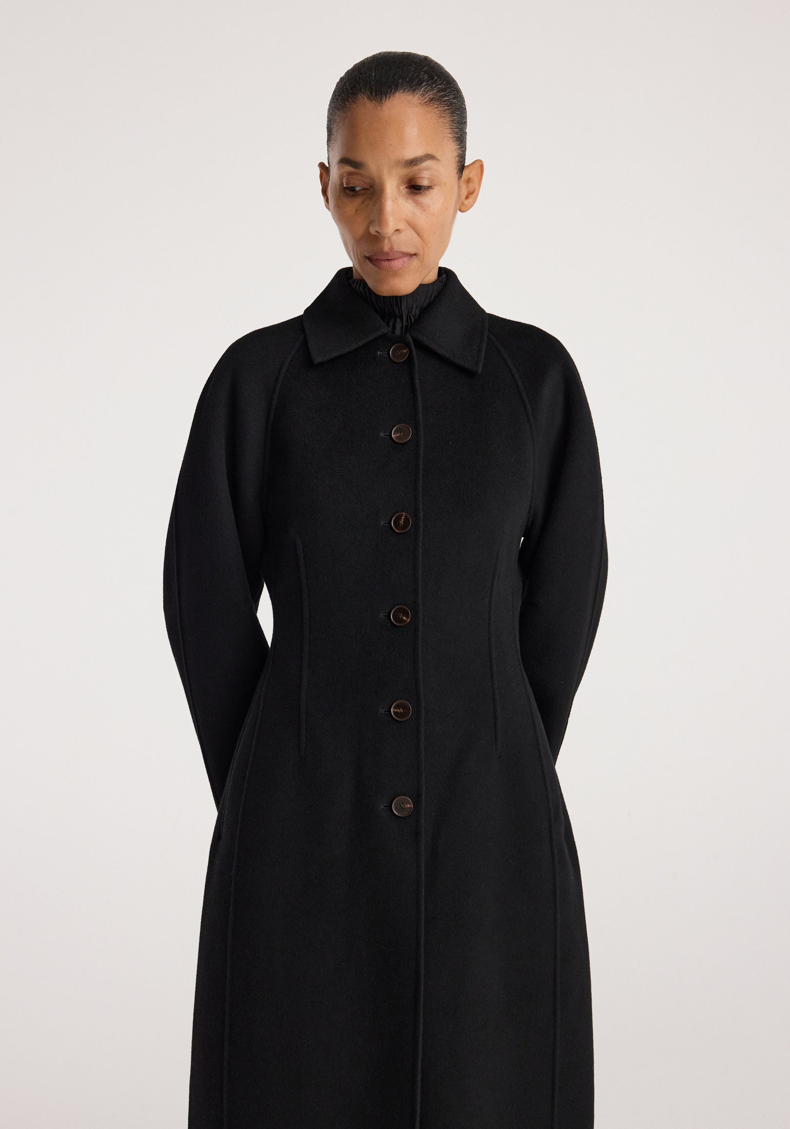 Woman wearing a sleek black Double-faced elongated sculptural coat | black with button closure and sculpted long sleeves against a plain light background.